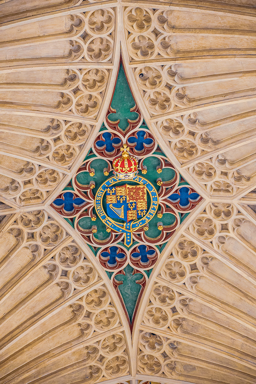 The image depicts an ornate ceiling design featuring a central coat of arms surrounded by intricate floral patterns and ribbed vaulting. The coat of arms includes heraldic symbols and text, and is encased in a circular frame with a green background and red and blue decorative elements. The surrounding ceiling is composed of stone ribs and recessed floral motifs, creating a visually striking and symmetrical pattern.