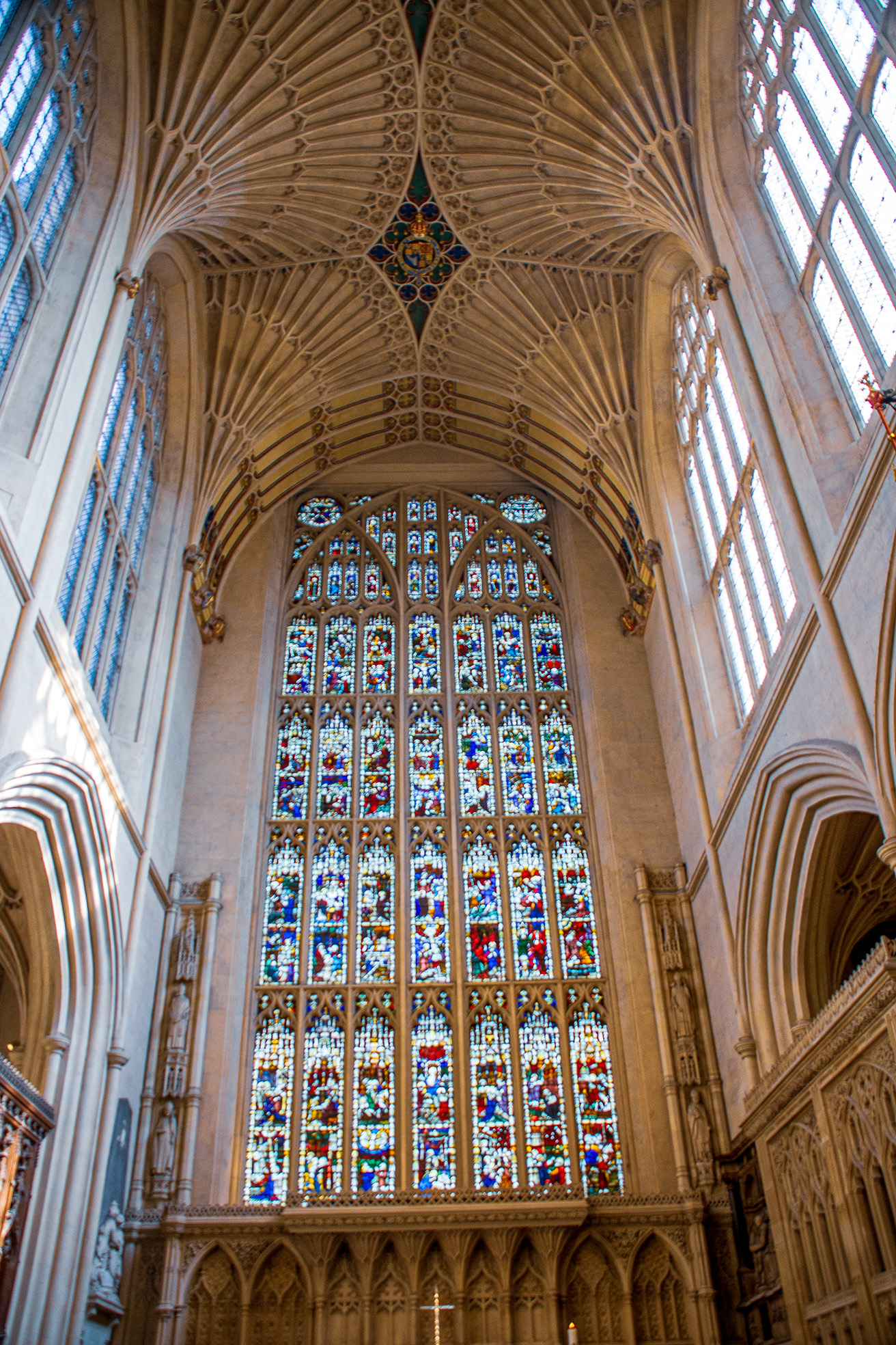 The image depicts the interior of a grand Gothic cathedral, showcasing intricate stone carvings, pointed arches, and a large, ornate stained glass window. The ceiling features a fan vault, a characteristic element of Gothic architecture, with detailed patterns and ribbed structures. The stained glass window is composed of numerous panels, each filled with vibrant, colorful depictions of religious scenes and figures. The overall atmosphere is one of reverence and historical significance, highlighting the architectural splendor of Gothic cathedrals.