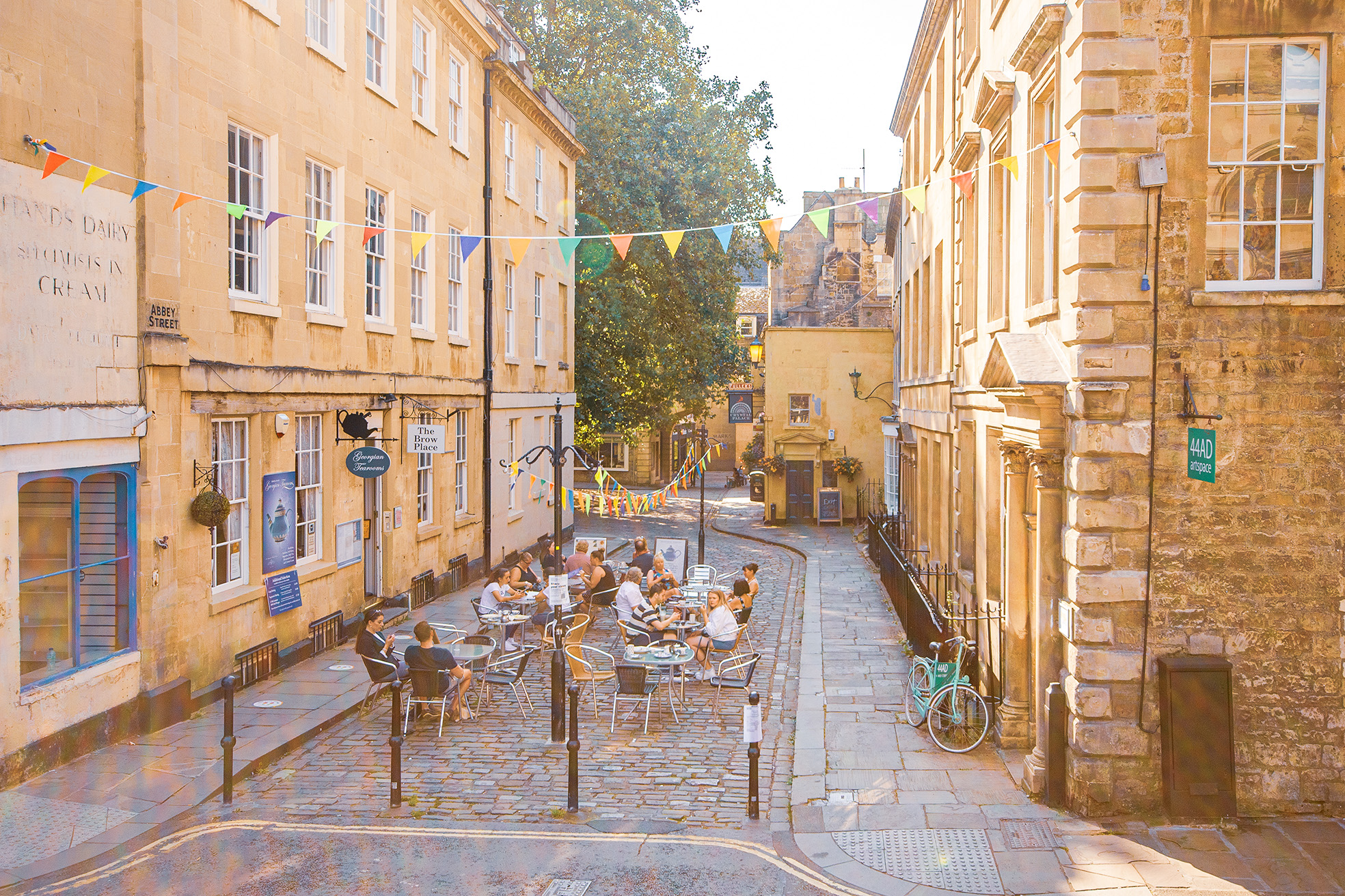 The image depicts a charming street scene in what appears to be a historic European town. The street is lined with old stone buildings, some with signs indicating businesses such as 'The Brownie Place' and 'Abbey Street.' There are colorful bunting flags strung across the street, adding a festive touch. People are seated at outdoor cafes, enjoying the pleasant weather. The street is narrow and cobblestoned, with a bicycle parked on the right side. The overall atmosphere is relaxed and inviting, suggesting a popular spot for locals and tourists alike.
