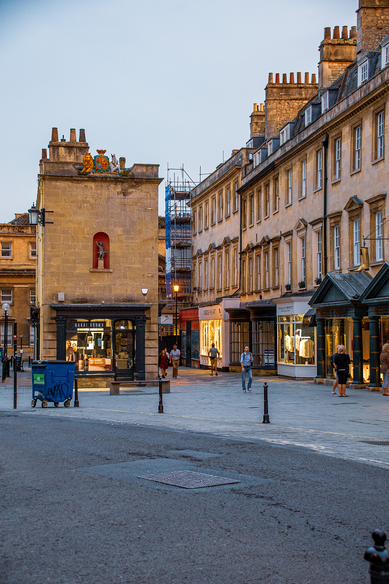 The image depicts a street scene in what appears to be a historic European city. The architecture is characterized by stone buildings with ornate details, including a prominent stone tower with a crest on top. The street is relatively quiet with a few pedestrians and some shops visible, including a jeweler and a clothing store. The overall atmosphere suggests a peaceful, early evening setting.