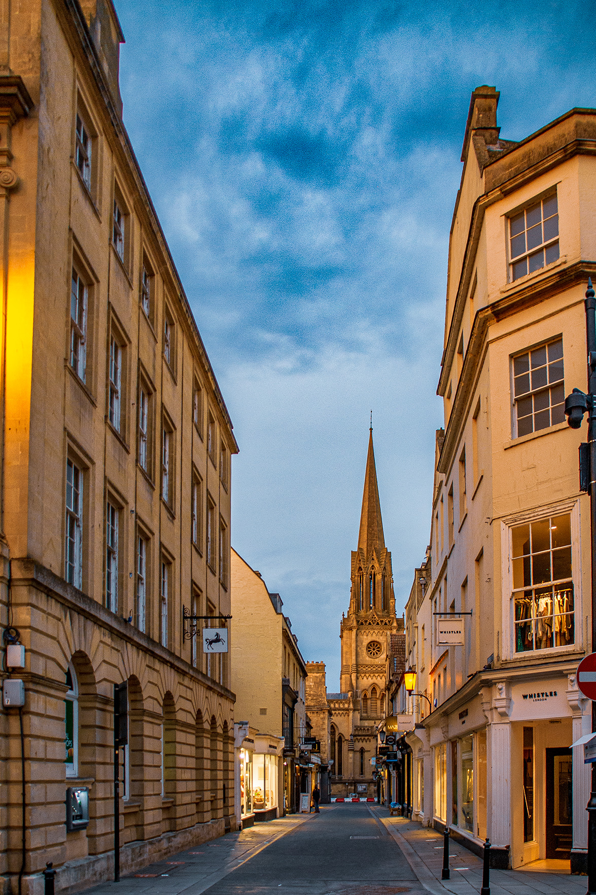 The image depicts a narrow street lined with buildings on either side, leading towards a large, ornate church with a tall spire in the distance. The buildings are a mix of modern and traditional architecture, with shops and businesses on the ground floors. The sky is clear with a hint of dusk, casting a warm glow on the scene. The street appears to be in a quiet, possibly historic, European town.