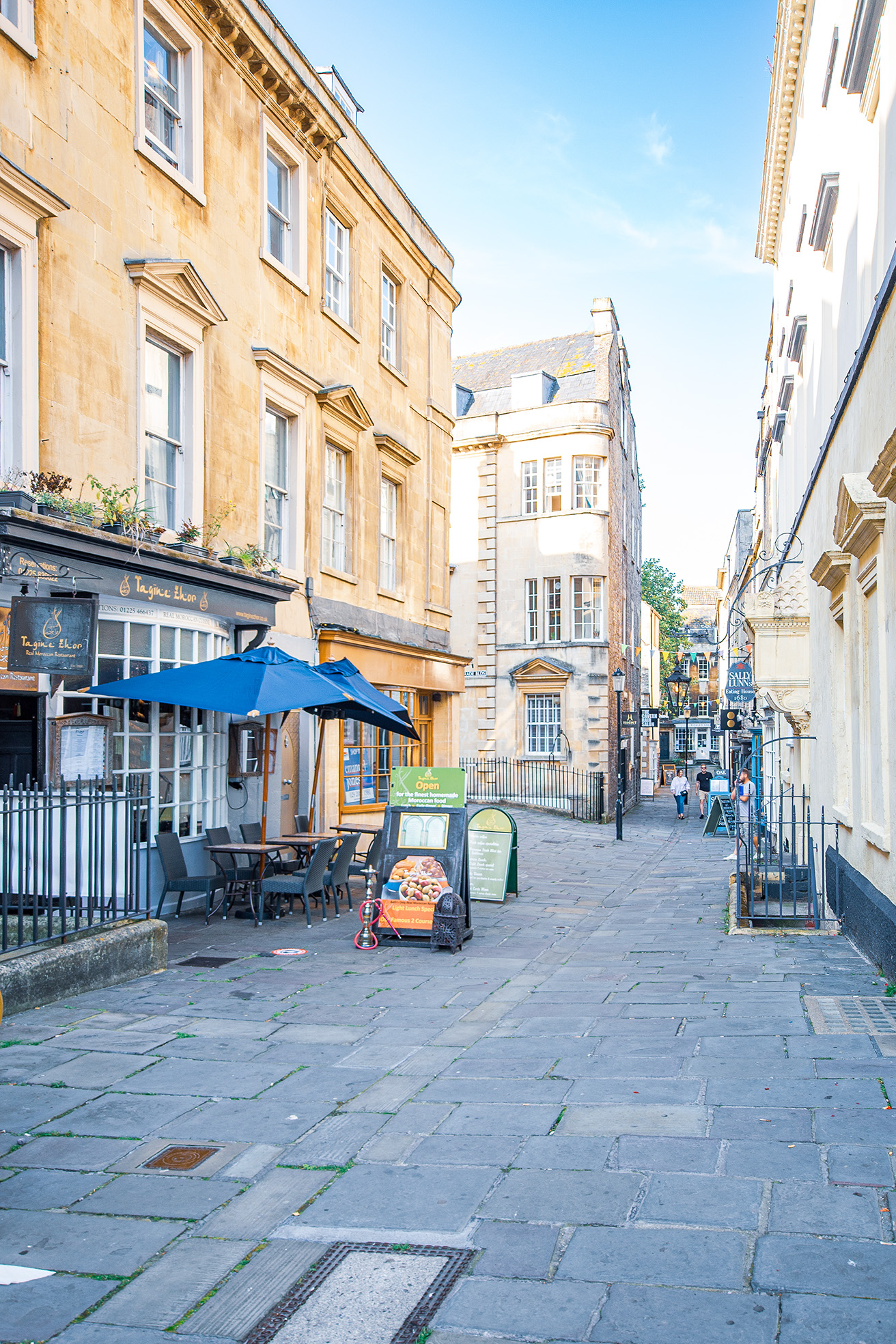 The image depicts a quaint, narrow street in what appears to be a historic European town. The street is lined with old, yellowish stone buildings featuring large windows and ornate architectural details. There are outdoor seating arrangements with tables and chairs under large umbrellas, suggesting the presence of cafes or restaurants. The street is paved with stone slabs and has a few pedestrians walking in the distance. The overall atmosphere is serene and inviting, with a clear blue sky overhead.