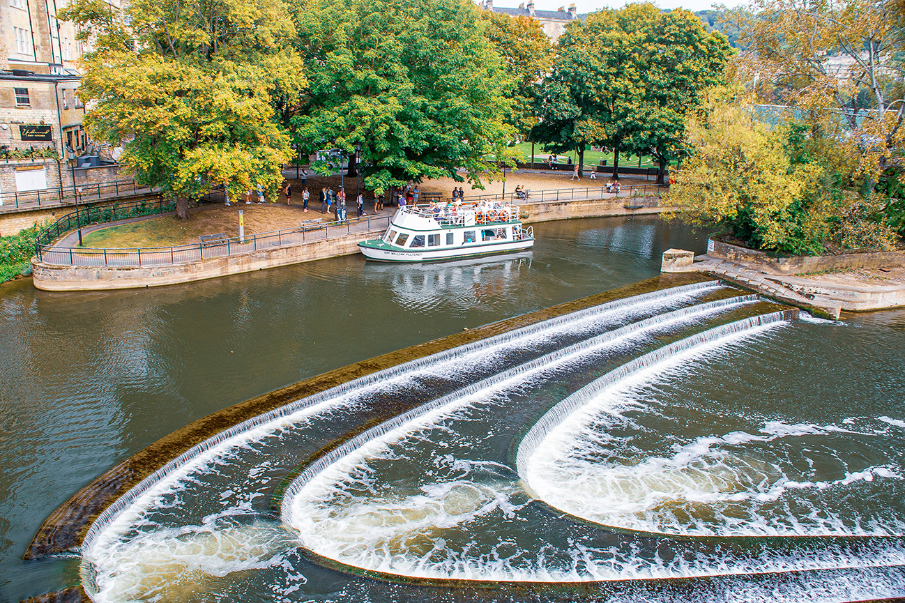 The image depicts a scenic view of a river with a weir and a boat. The weir has a series of cascades, creating a picturesque effect. On the left side, there is a lush, green park with trees displaying autumn foliage. People are walking along the path in the park, and a boat is seen on the river, carrying passengers. The backdrop includes buildings and a clear sky, suggesting a pleasant day.