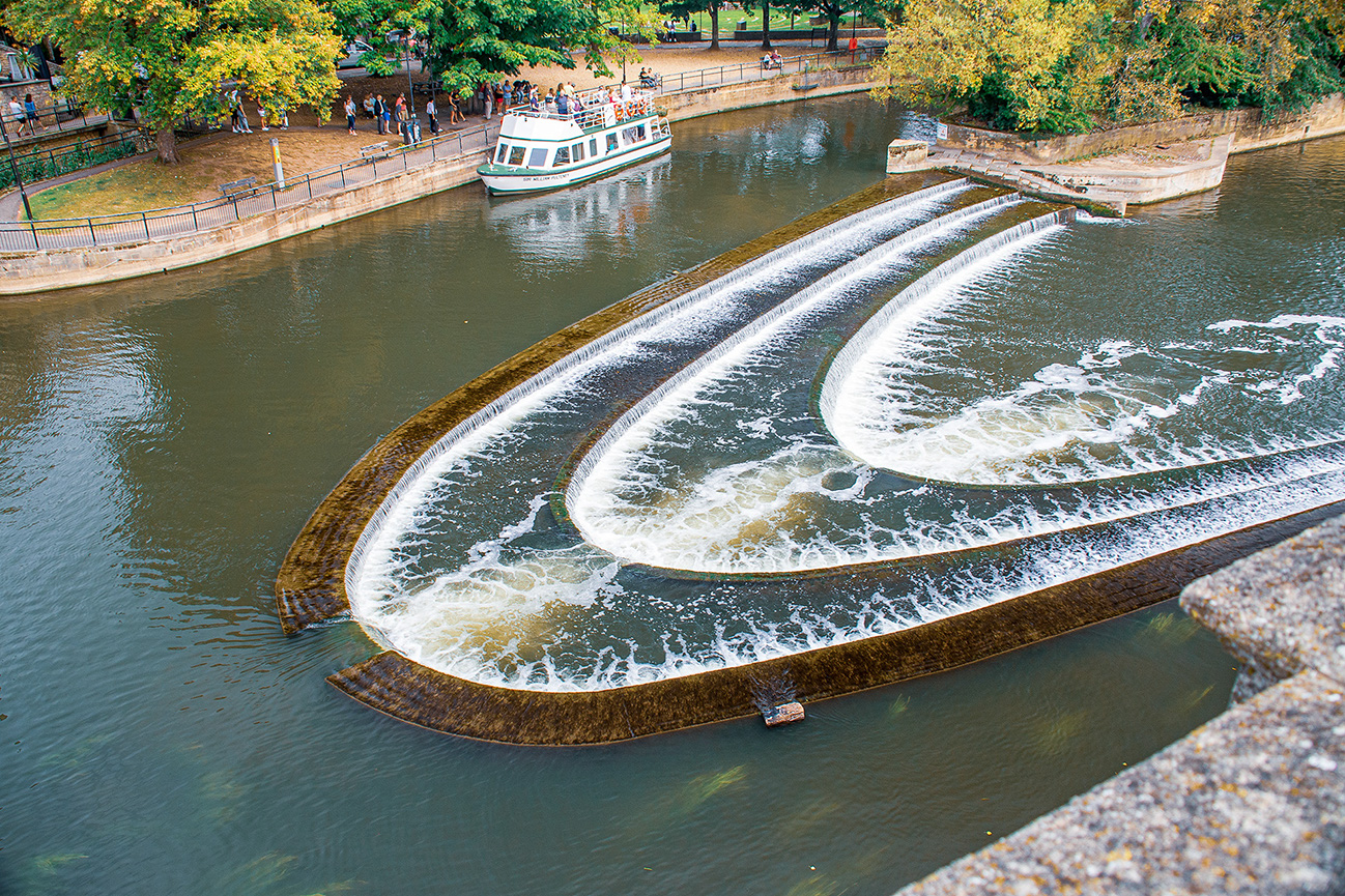 The image depicts a scenic view of a canal with a series of cascading water steps, creating a visually appealing effect. In the background, there is a boat docked near a pathway lined with trees and people walking along the edge of the canal. The setting appears to be a park or a recreational area, with clear, calm water and a serene atmosphere.