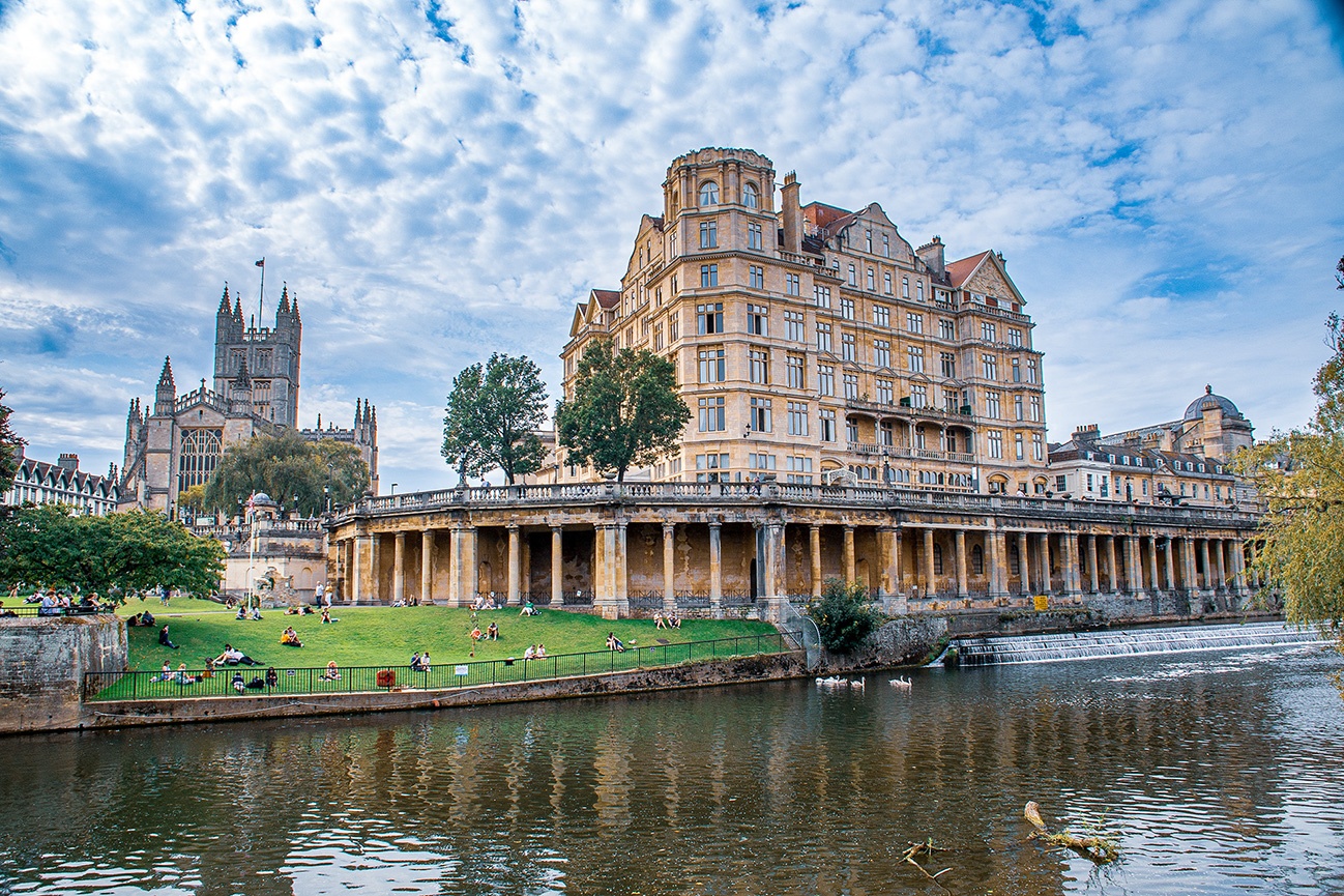 The image depicts a picturesque scene of a park in an academic setting, likely a university. The park features a large grassy area with numerous people relaxing, sitting, and enjoying the outdoors. In the background, there are historic buildings with classic architectural styles, including towers and ornate facades. The scene is framed by trees and a body of water in the foreground, adding to the serene atmosphere. The overall setting suggests a blend of natural beauty and historical architecture, creating a tranquil and inviting environment.