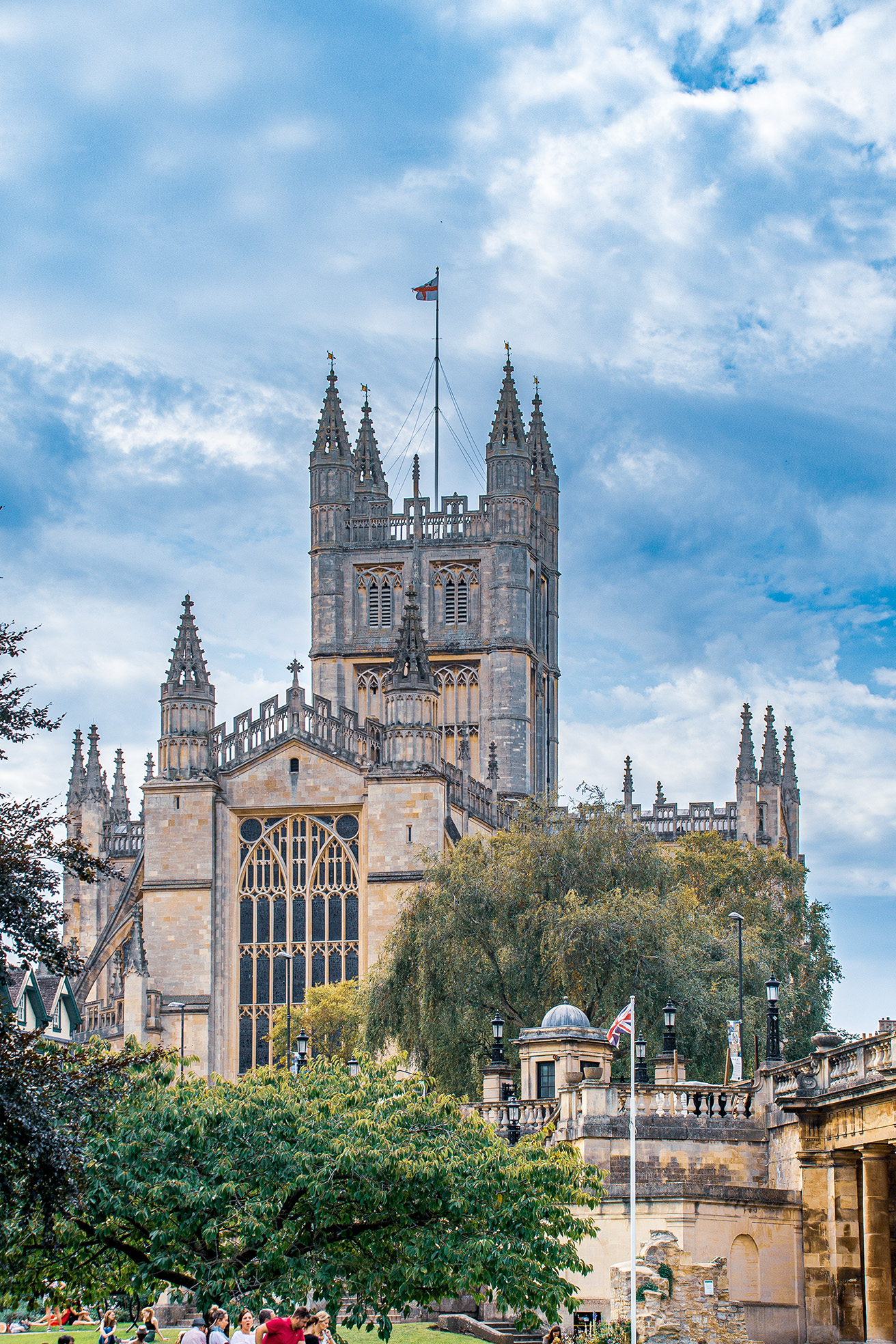 The image depicts a historic and grand Gothic-style building, likely a cathedral or university, with intricate stonework and large windows. The structure features multiple spires and a central tower with a flag flying at the top. The building is surrounded by lush greenery, including trees and shrubs, with people visible in the foreground enjoying the outdoor space. The sky above is partly cloudy, adding to the serene atmosphere.