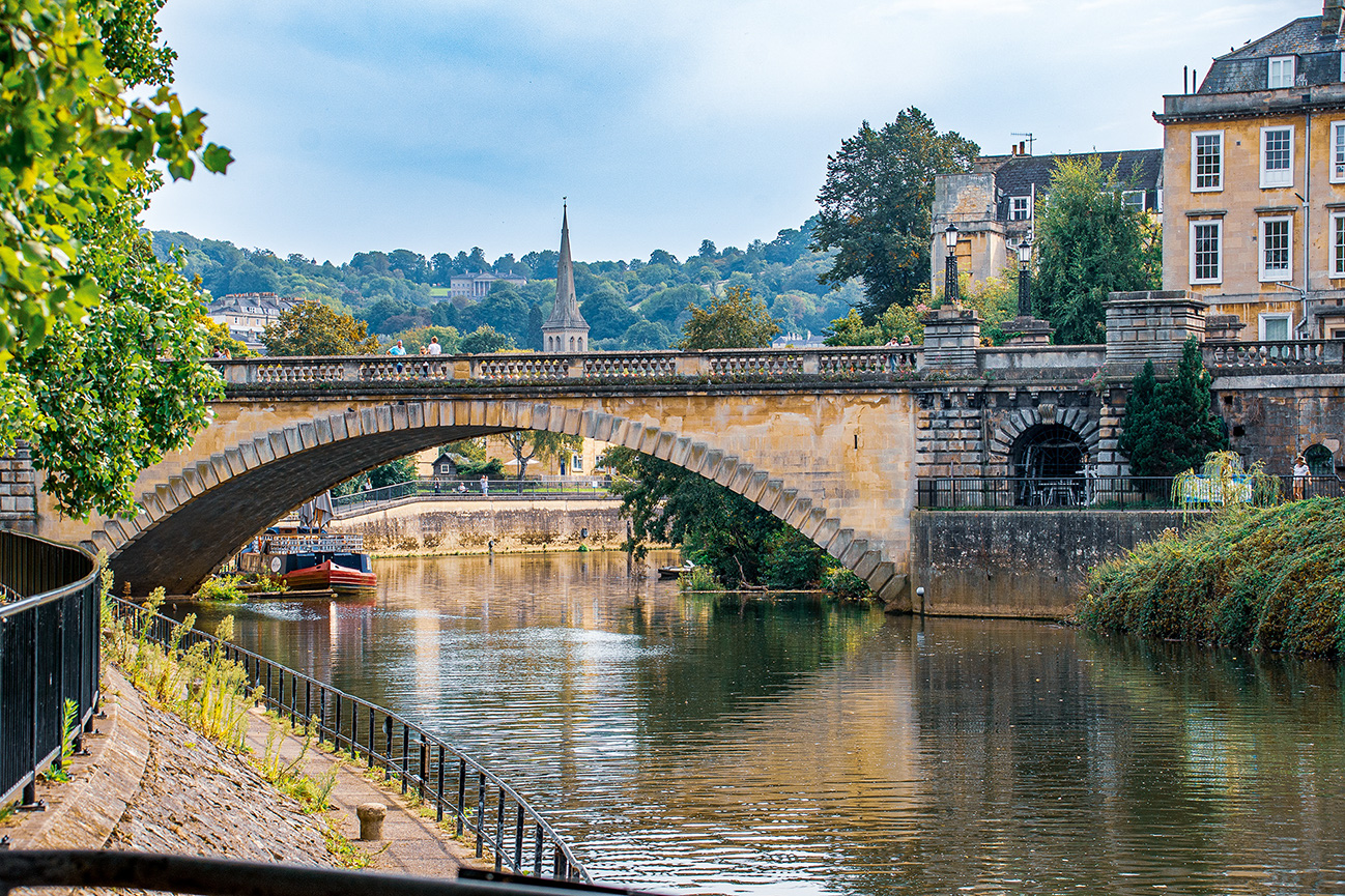 The image depicts a scenic view of a historic stone bridge arching over a calm river. The bridge is adorned with decorative balustrades and is flanked by stone buildings on either side. In the background, a spire rises above the tree line, adding to the picturesque setting. The river below reflects the bridge and surrounding greenery, creating a serene and picturesque scene. The area appears to be a blend of natural beauty and architectural elegance, likely in a quaint, historic town.