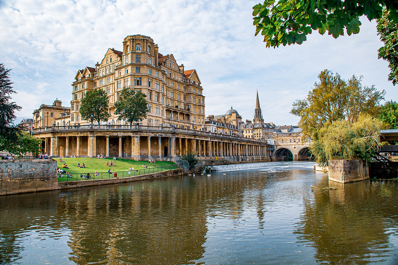 The image depicts a scenic view of the Pulteney Bridge and the historic buildings along the River Avon in Bath, England. The Pulteney Bridge, notable for its shops and historic architecture, spans the river with a series of columns. On the left side of the image, people are seen relaxing on the grassy area near the river. The buildings exhibit classic Georgian architecture, with large windows and ornate details. In the background, the spire of Bath Abbey is visible, adding to the historic charm of the scene. The overall atmosphere is serene and picturesque, capturing the essence of this iconic location.