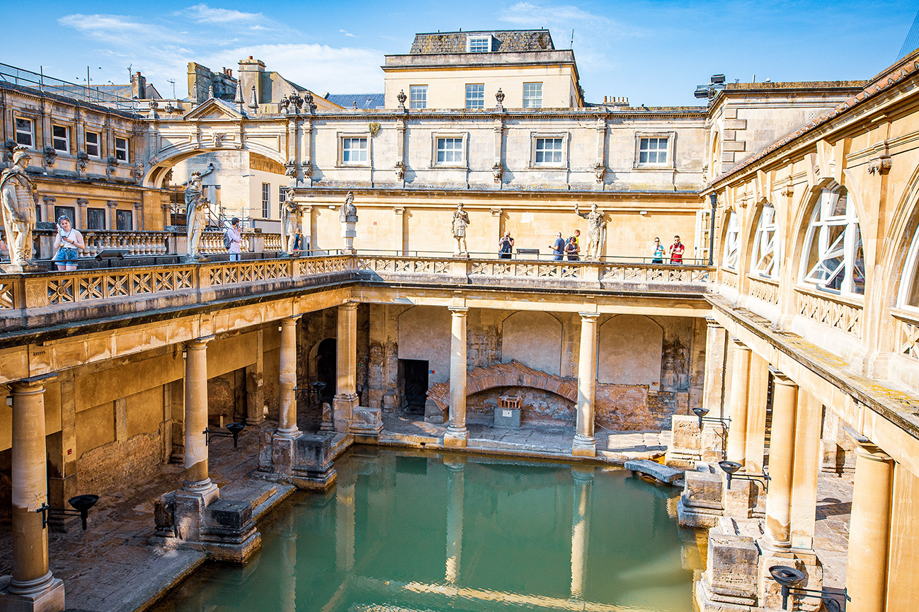 The image depicts the ancient Roman Baths in Bath, England. The scene shows a historic, open-air pool surrounded by classical Roman architecture, including columns and statues. The upper level features a terrace with people walking and observing the view. The buildings are constructed with light-colored stone, and the overall atmosphere is one of historical significance and architectural grandeur.