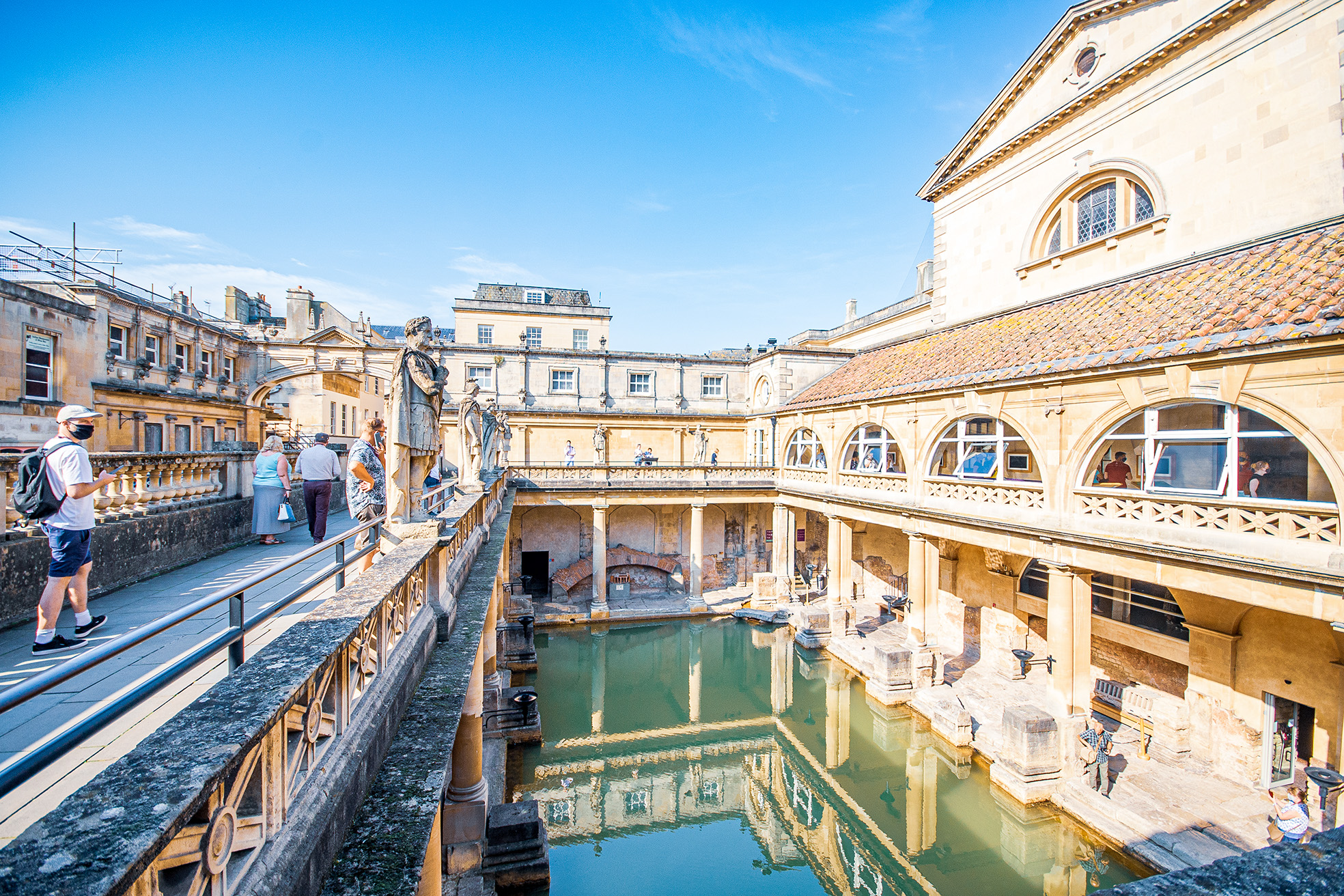 The image depicts the ancient Roman Baths in Bath, England, showcasing the historic site's architectural beauty and the pool of water in the center. Visitors can be seen walking around the site, observing the well-preserved ruins and the surrounding structures.