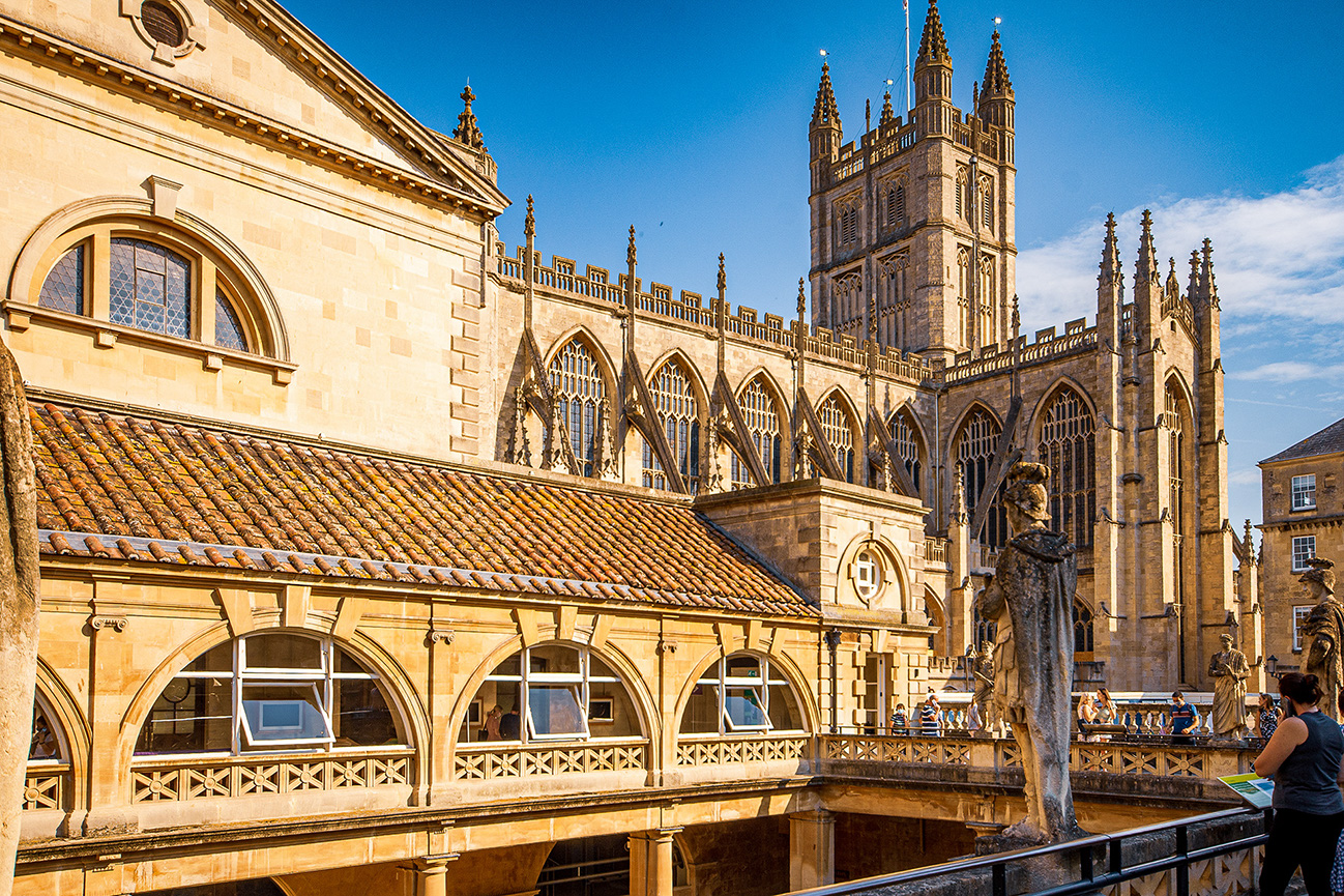 The image depicts a historic architectural structure, likely a cathedral or a large church, characterized by its Gothic style. The building features pointed arches, ribbed vaults, and flying buttresses. In the foreground, there is a bridge with arches and a statue of a person riding a horse. The scene is bustling with people, suggesting it is a popular tourist destiUK. The sky is clear and blue, indicating good weather.