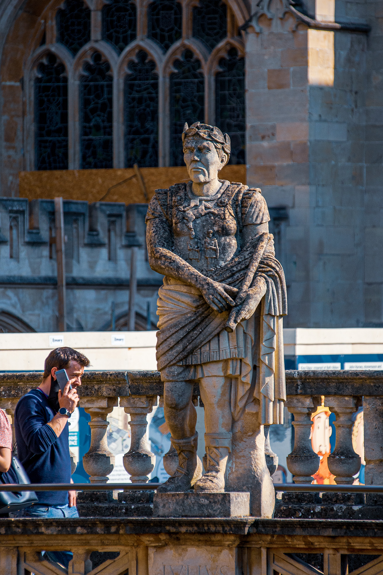 The image depicts a stone statue of a historical figure, possibly a knight or a king, standing in front of a Gothic-style building with intricate window designs. The statue is detailed, showing the figure in armor with a cloak draped over the shoulders. In the foreground, a man is seen talking on a mobile phone, leaning against a stone balustrade. The scene suggests a tourist Bath, likely a historical or cultural site.