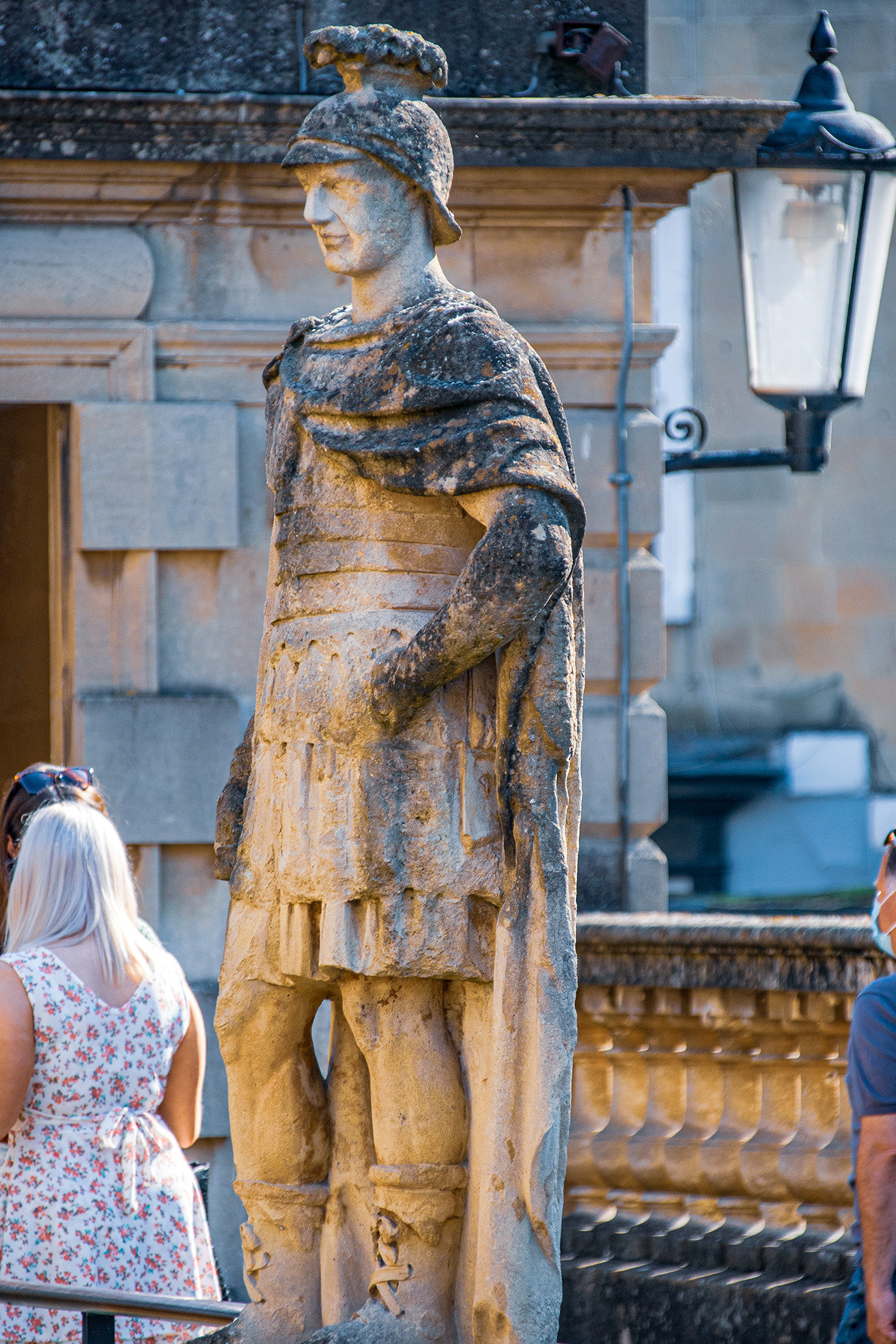 The image depicts a weathered stone statue of a Roman soldier wearing a helmet and armor, standing in front of a classical building with stone walls and a lamp post. The statue appears to be part of a historical or cultural site, with people observing and taking photos of it.