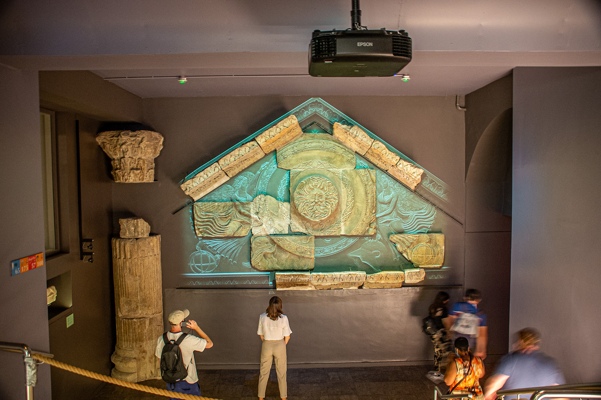 The image depicts a museum exhibit featuring an ancient, intricately designed artifact displayed on a wall. The artifact appears to be a reconstructed piece, possibly a frieze or a section of a larger structure, with detailed carvings and a central circular motif. Several visitors are observing the exhibit, with some taking photographs. The setting includes other ancient artifacts and a projector mounted on the ceiling.