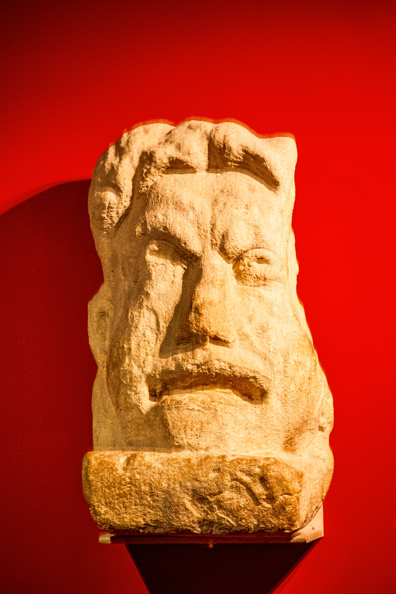 The image depicts an ancient stone sculpture of a bearded face with a serious expression, set against a red background. The sculpture appears to be of a man with detailed facial features, including prominent eyebrows, a strong nose, and a beard. The stone used is light-colored, and the sculpture is displayed on a pedestal. The red background enhances the contrast, highlighting the intricate details of the stone carving.