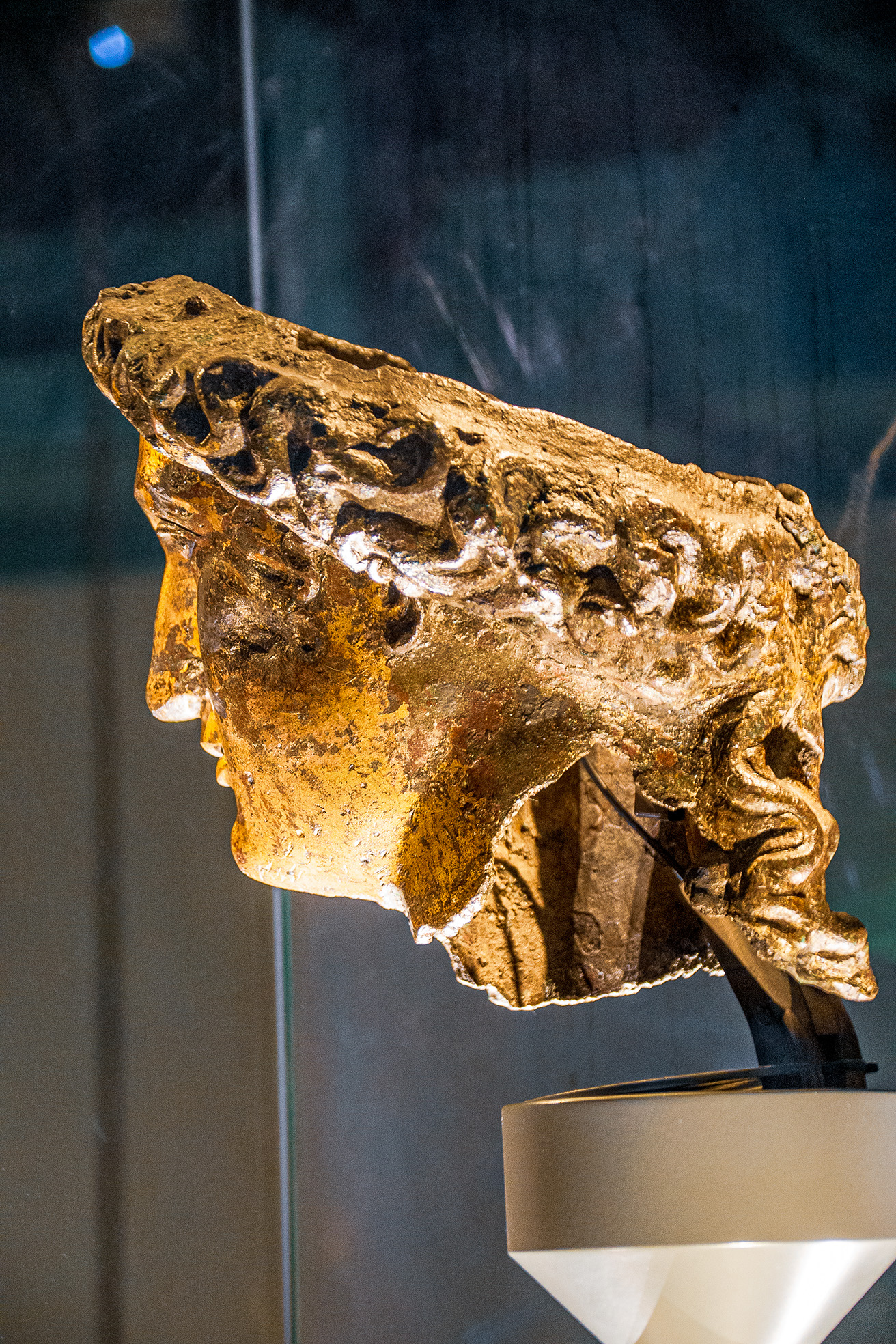 The image shows a statue of a head displayed in a museum setting. The statue is mounted on a stand and is illuminated by a spotlight from below, highlighting its detailed structure and texture.