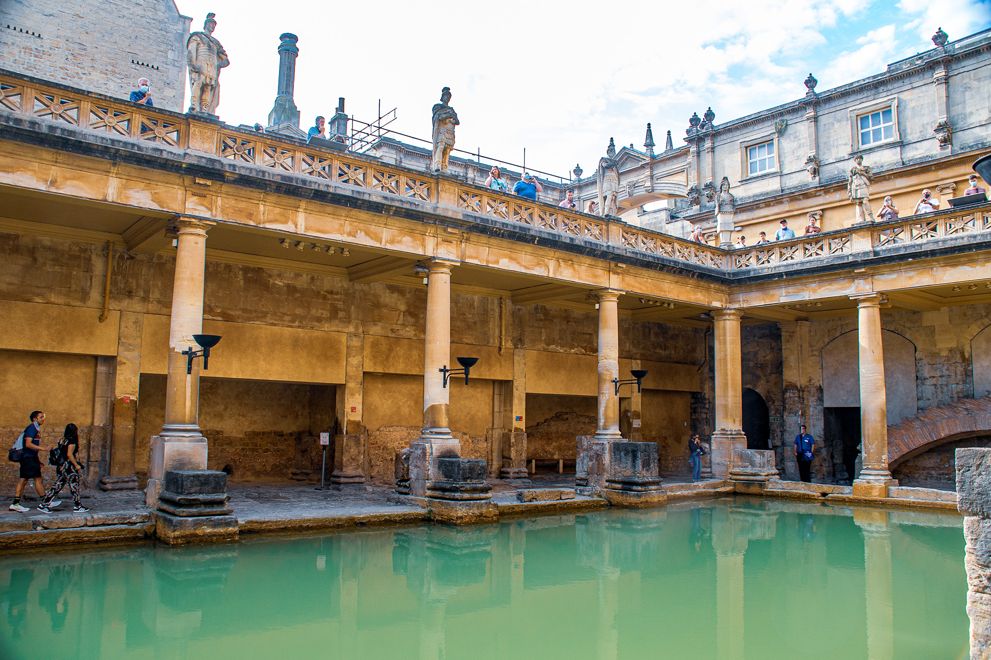 The image depicts the ancient Roman Baths in Bath, England. The scene shows a historic site with a large, greenish pool of water in the foreground, surrounded by stone columns and arches. Above the columns, there is a terrace with statues and people observing the view. The architecture is classical, with intricate details and a mix of stone and brick materials. The sky is partly cloudy, adding to the historic ambiance of the Bath.