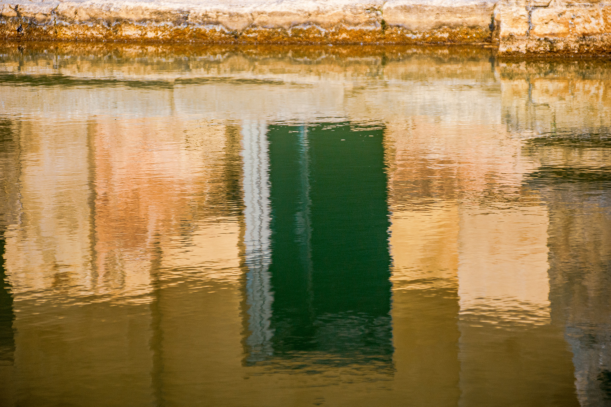 The image shows a close-up view of a rusty metal surface, possibly part of an old structure or machinery, with a reflective water surface below. The water reflects the rusty texture and colors of the metal, creating a distorted mirror image. The overall scene conveys a sense of decay and stillness, with the interplay of natural and man-made elements.