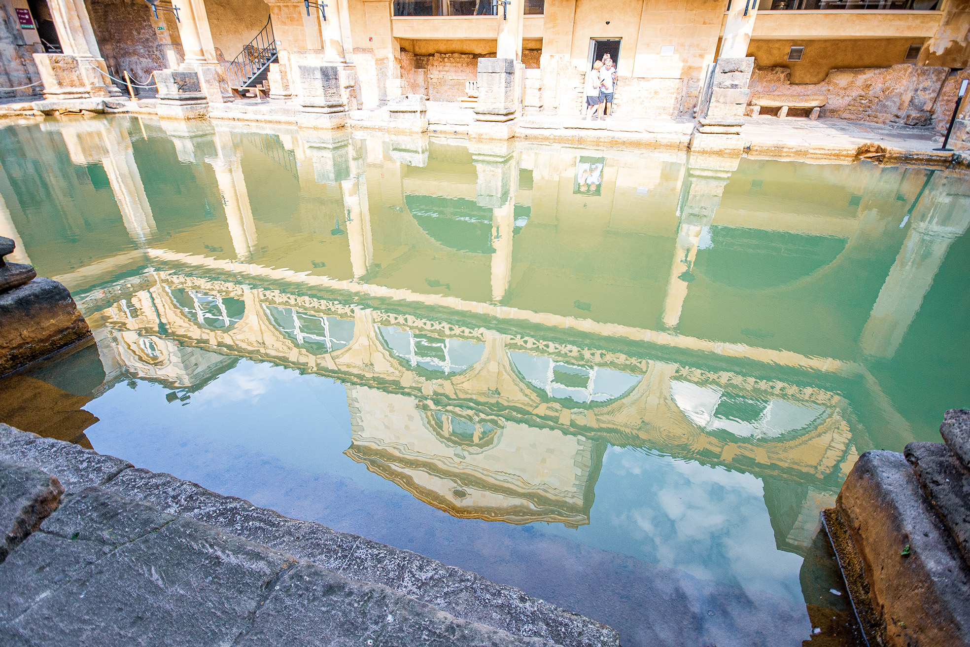 The image depicts an ancient Roman bathhouse, featuring a large, rectangular pool of water surrounded by stone columns and architectural ruins. The water in the pool is greenish and reflects the surrounding structures. Two people are standing near the edge of the pool, observing the site. The overall scene suggests a historical and archaeological setting, likely a popular tourist attraction.