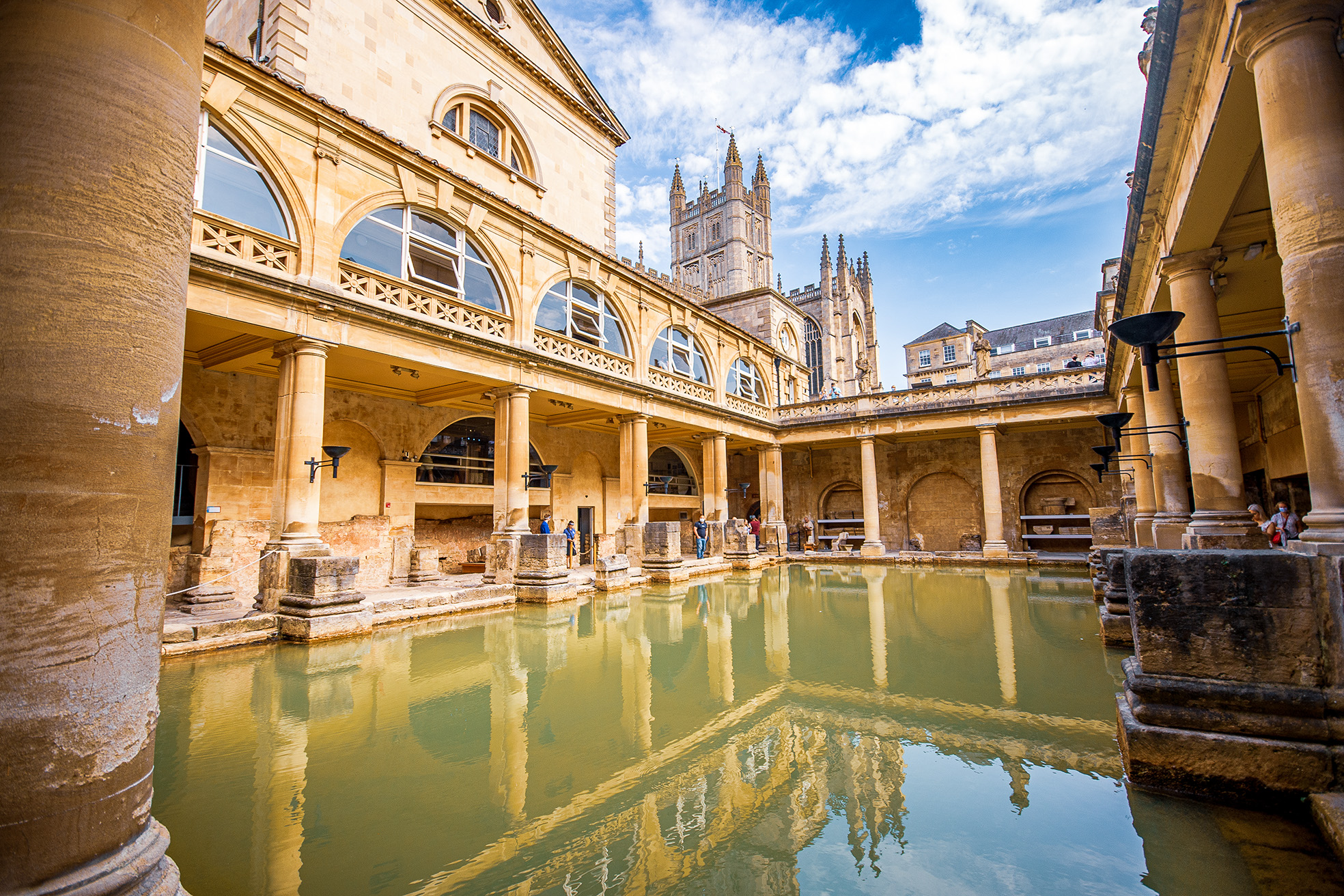 The image depicts the ancient Roman Baths in Bath, England. The scene shows a historic, open-air pool surrounded by stone columns and arches. The architecture is Roman in style, with a mix of ancient and medieval elements. The sky is partly cloudy, and the water in the pool is greenish, reflecting the surrounding structures. In the background, a Gothic-style cathedral tower is visible.