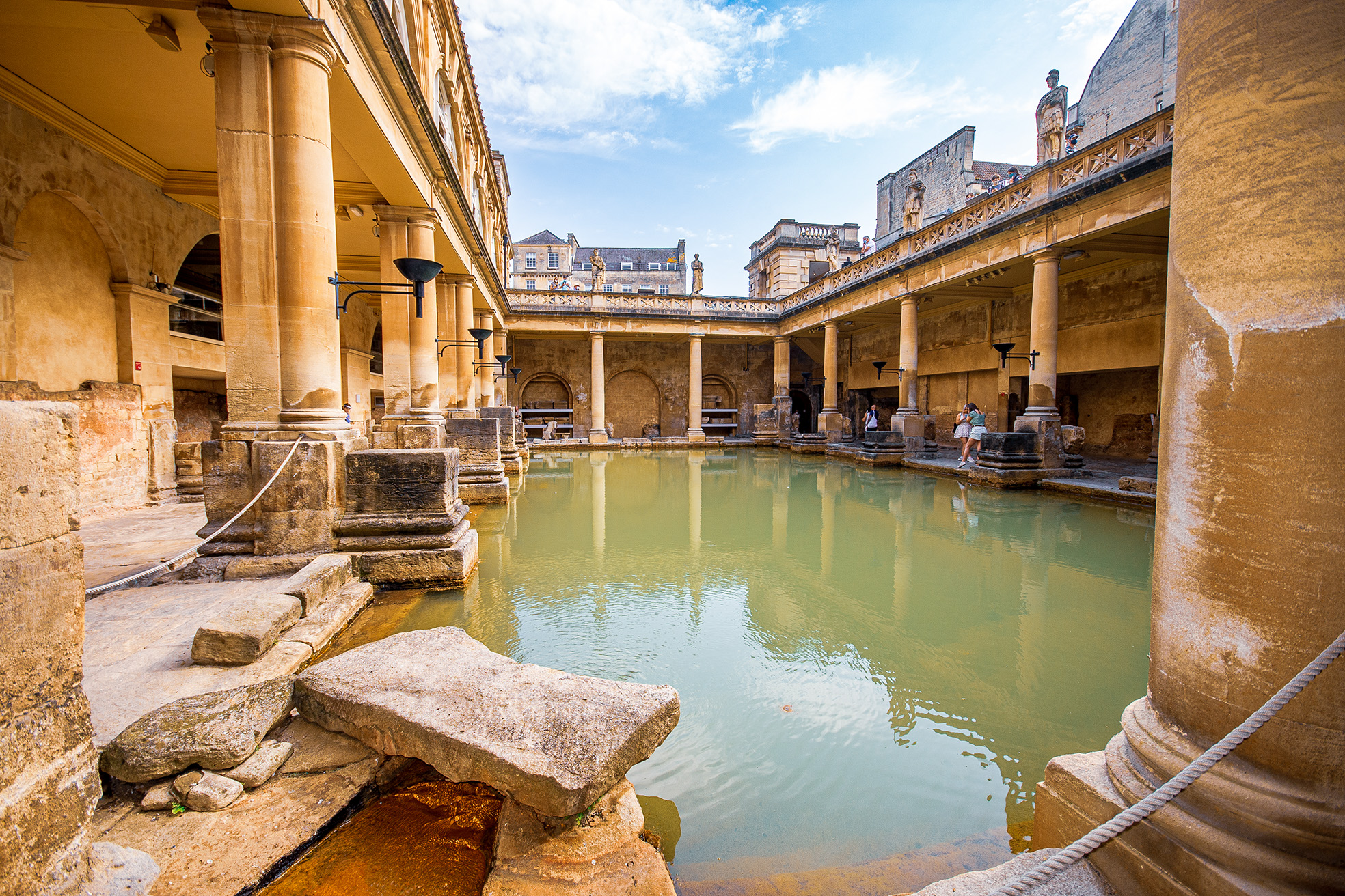 The image depicts the ancient Roman Baths in Bath, England. The scene shows a large, open-air pool surrounded by historic stone architecture, including columns and statues. The water in the pool is a greenish color, and the area is framed by weathered stone structures. A few people can be seen walking along the edges of the pool, and the sky above is partly cloudy. The overall atmosphere is one of historical significance and architectural grandeur.
