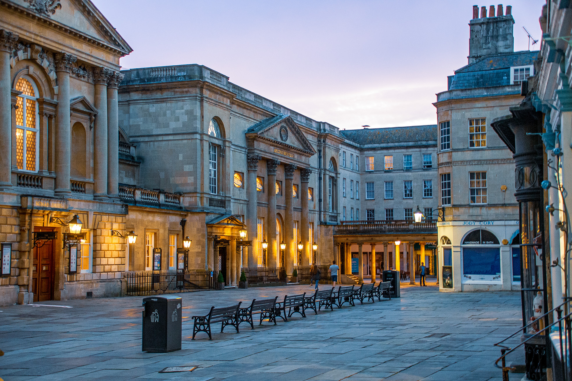 The image depicts an architectural scene of a courtyard or plaza surrounded by historic buildings with classical architectural elements such as columns, arches, and pediments. The buildings are illuminated by street lamps, creating a warm and inviting atmosphere. The courtyard features several benches and trash bins, and a few people can be seen walking or sitting, suggesting a public space likely located in a European city.