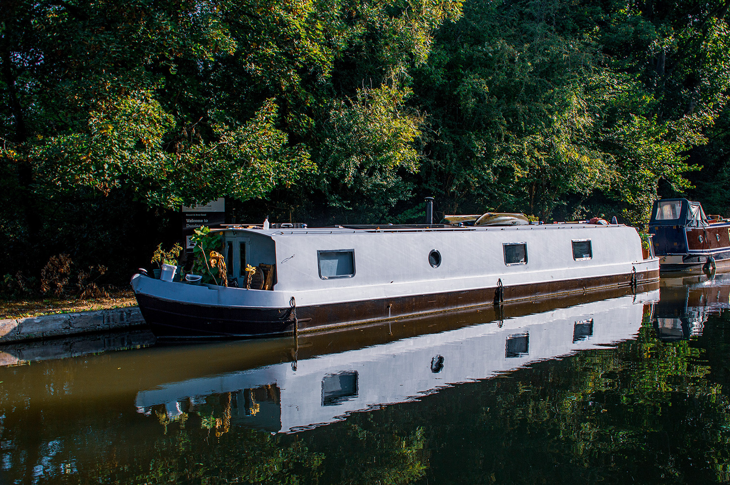 The image depicts a serene scene of a narrowboat moored on a calm canal, surrounded by lush greenery and trees. The boat is white with several windows and a round porthole, and it is equipped with various plants and items on the roof. The water is still, creating a perfect reflection of the boat and the surrounding environment. Another boat is partially visible to the right. The setting suggests a peaceful, rural Brassknocker, ideal for a tranquil boating experience.