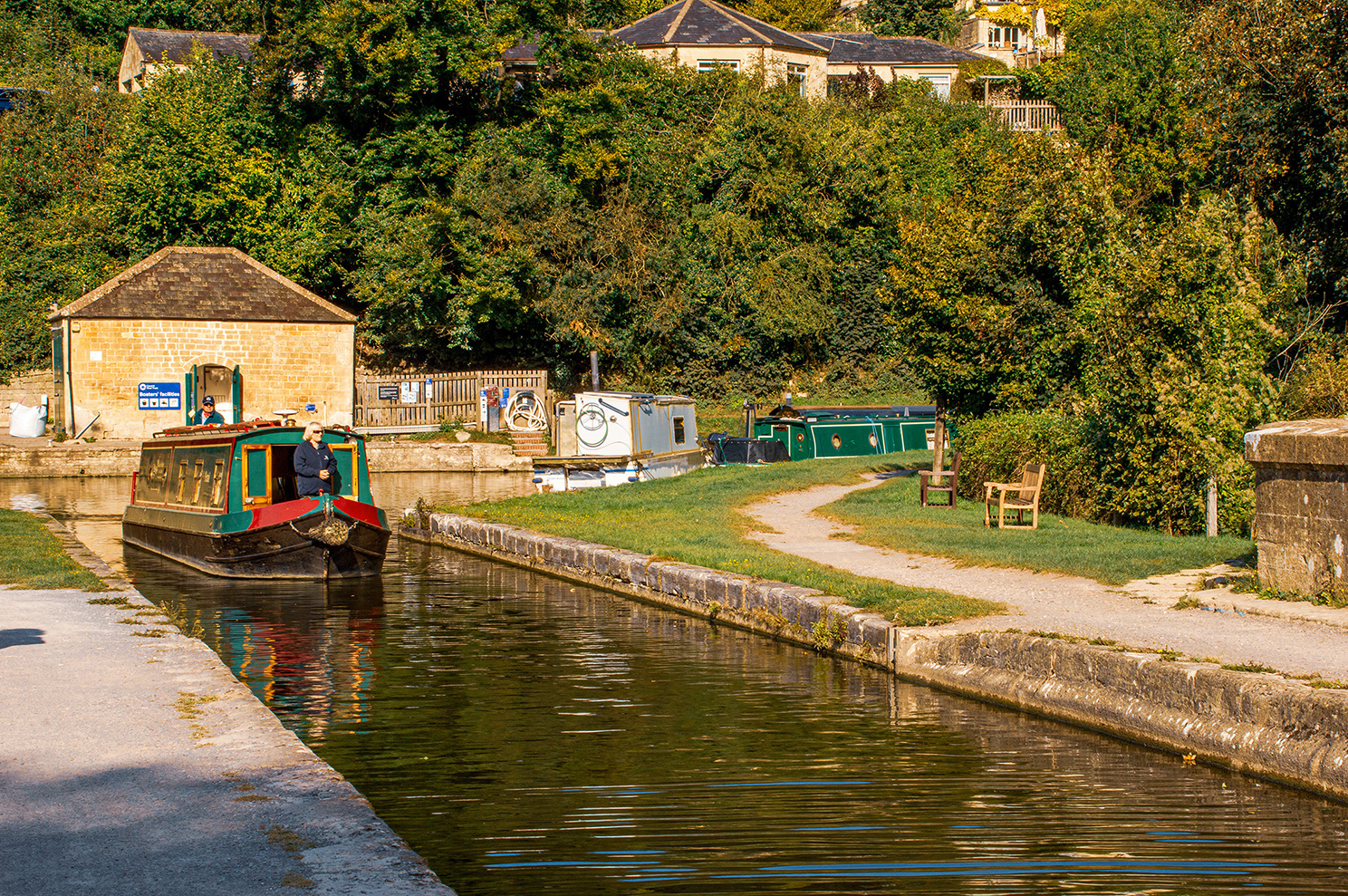The image depicts a serene canal scene with narrowboats moored along the side. A man is steering a narrowboat towards the camera. The canal is lined with stone walls and surrounded by lush greenery. There are several narrowboats docked on the opposite side, and a small stone building with a sign is visible. The overall atmosphere is peaceful and picturesque, suggesting a quiet, rural setting.