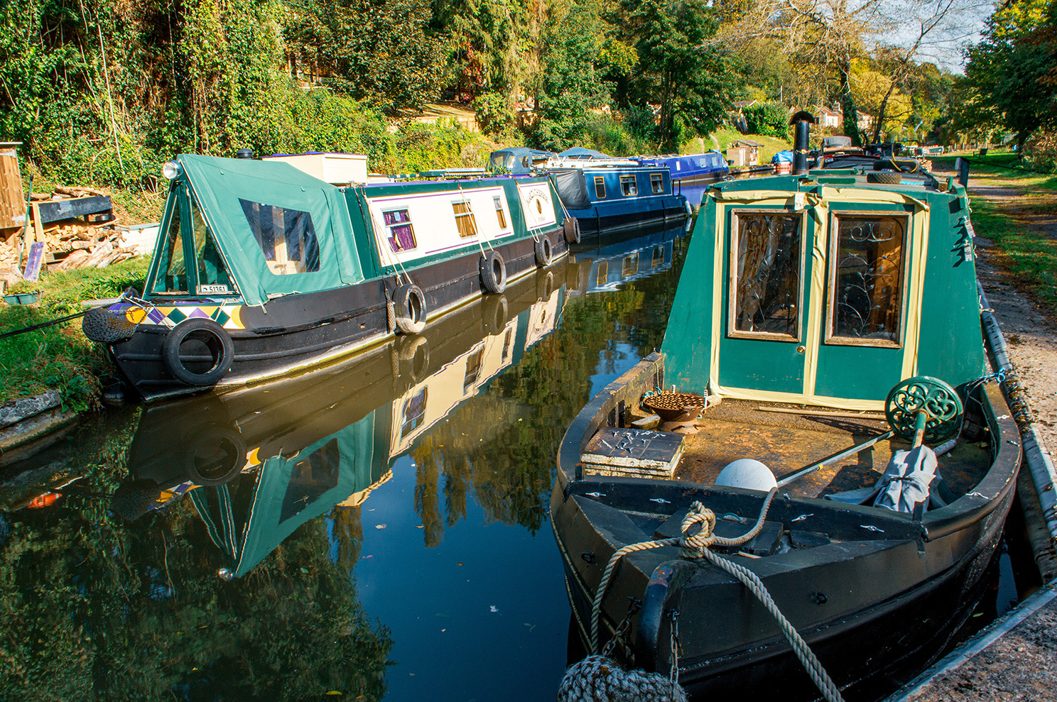 The image shows a serene canal scene with several narrowboats moored along the bank. The boats are painted in various colors, with the foreground featuring a green narrowboat with a wooden deck. The surrounding area is lush with greenery, and the water in the canal is calm, reflecting the boats and trees. The scene conveys a peaceful, rural atmosphere, typical of narrowboat moorings in the countryside.