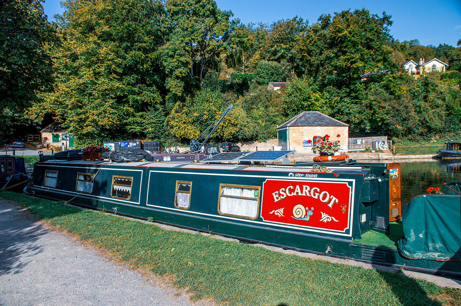 The image depicts a narrowboat named 'Escargot' moored on a canal. The boat is painted green with a red and white sign featuring snails. It is adorned with potted plants and has various equipment on its roof. The surroundings include a grassy path, trees, and a few buildings in the background.
