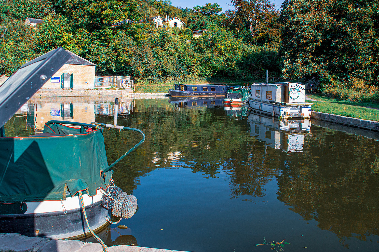 The image depicts a serene canal scene with several narrowboats moored along the bank. The water is calm, reflecting the boats and surrounding greenery. The scene is framed by the bow of a boat in the foreground, with a green canopy and a fender hanging off the side. The background features lush trees and a few buildings, including a small stone structure with green shutters. The overall atmosphere is peaceful and picturesque, typical of a quiet canal setting.