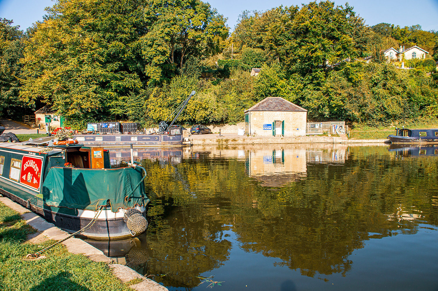 The image depicts a serene canal scene with several narrowboats moored along the bank. The water is calm, reflecting the surrounding greenery and boats. There is a small stone building with a green door and a blue sign, likely a canal office or facility. The area is lush with trees and foliage, indicating a peaceful, rural setting. The overall atmosphere is tranquil and picturesque.