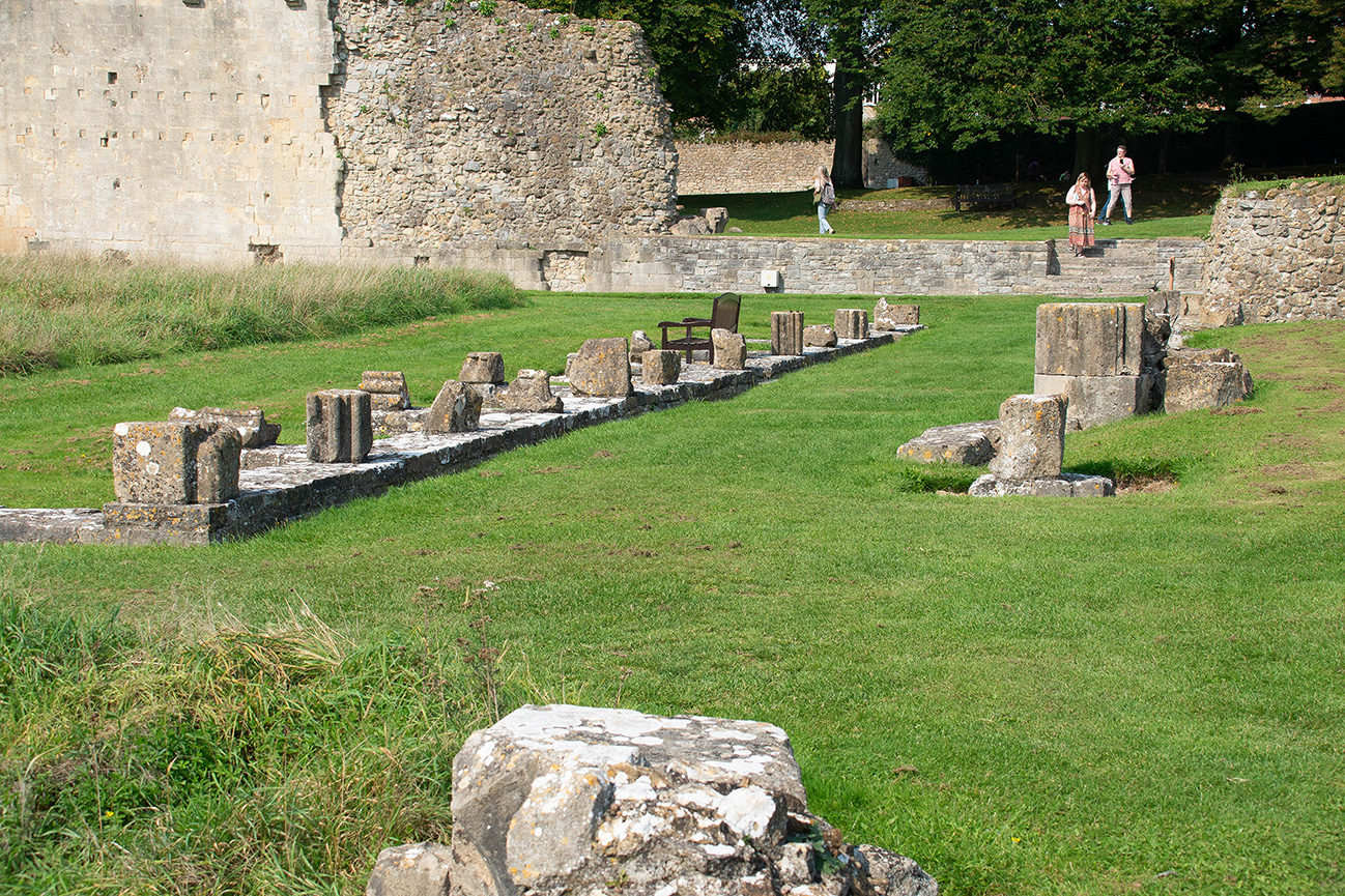 The image depicts an ancient, partially ruined structure, possibly a historical site or archaeological ruin. The remains consist of stone foundations and scattered stones, suggesting the outlines of former walls or buildings. The area is grassy and well-maintained, with a few people walking around, indicating it might be a tourist attraction or a preserved historical Brassknocker. The backdrop includes old stone walls and lush greenery, enhancing the historical ambiance of the scene.