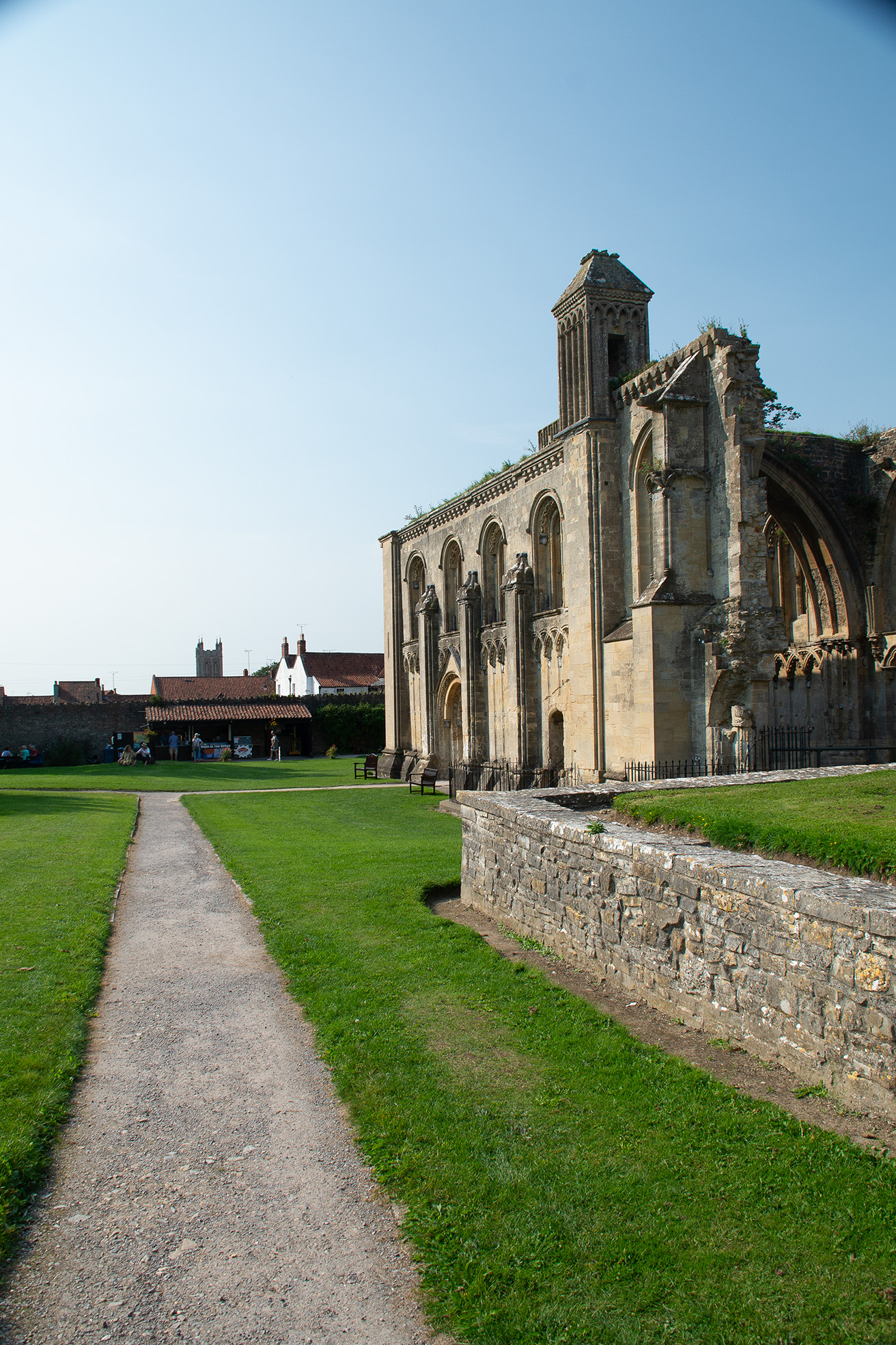 The image depicts the ruins of an old stone building, likely a historical or medieval structure, situated in a well-maintained grassy area. The building features large, arched windows and a tower-like structure on the right side. A gravel path leads up to the building, and there are a few people visible in the background, suggesting it might be a tourist site or a place of historical significance.