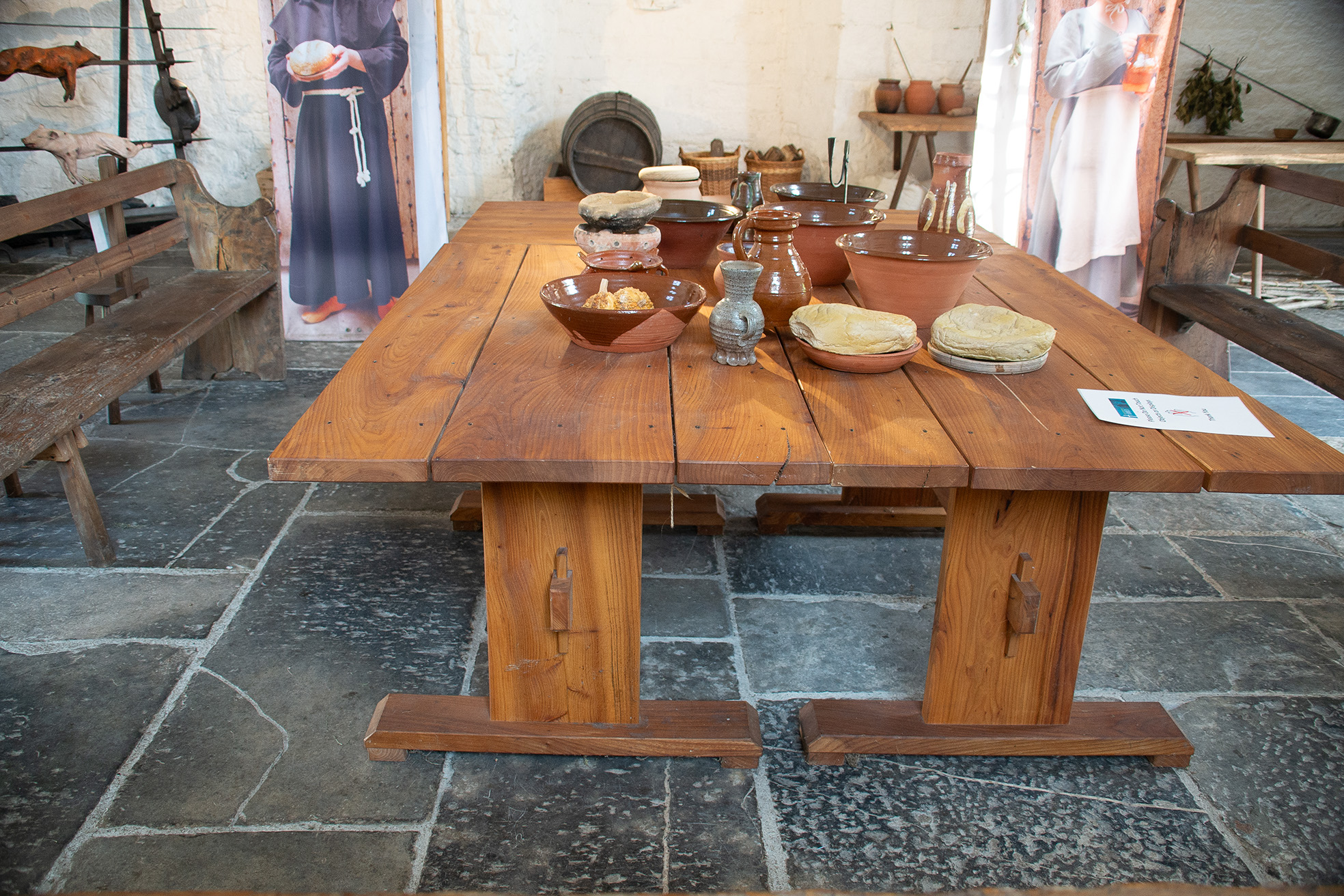 The image depicts a rustic, medieval-style dining setup with a wooden table and benches. The table is set with various pottery dishes, bowls, and jugs, some containing food items. The room features stone walls and floor, with a few decorative elements such as posters and hanging items, suggesting a historical or reenactment setting.