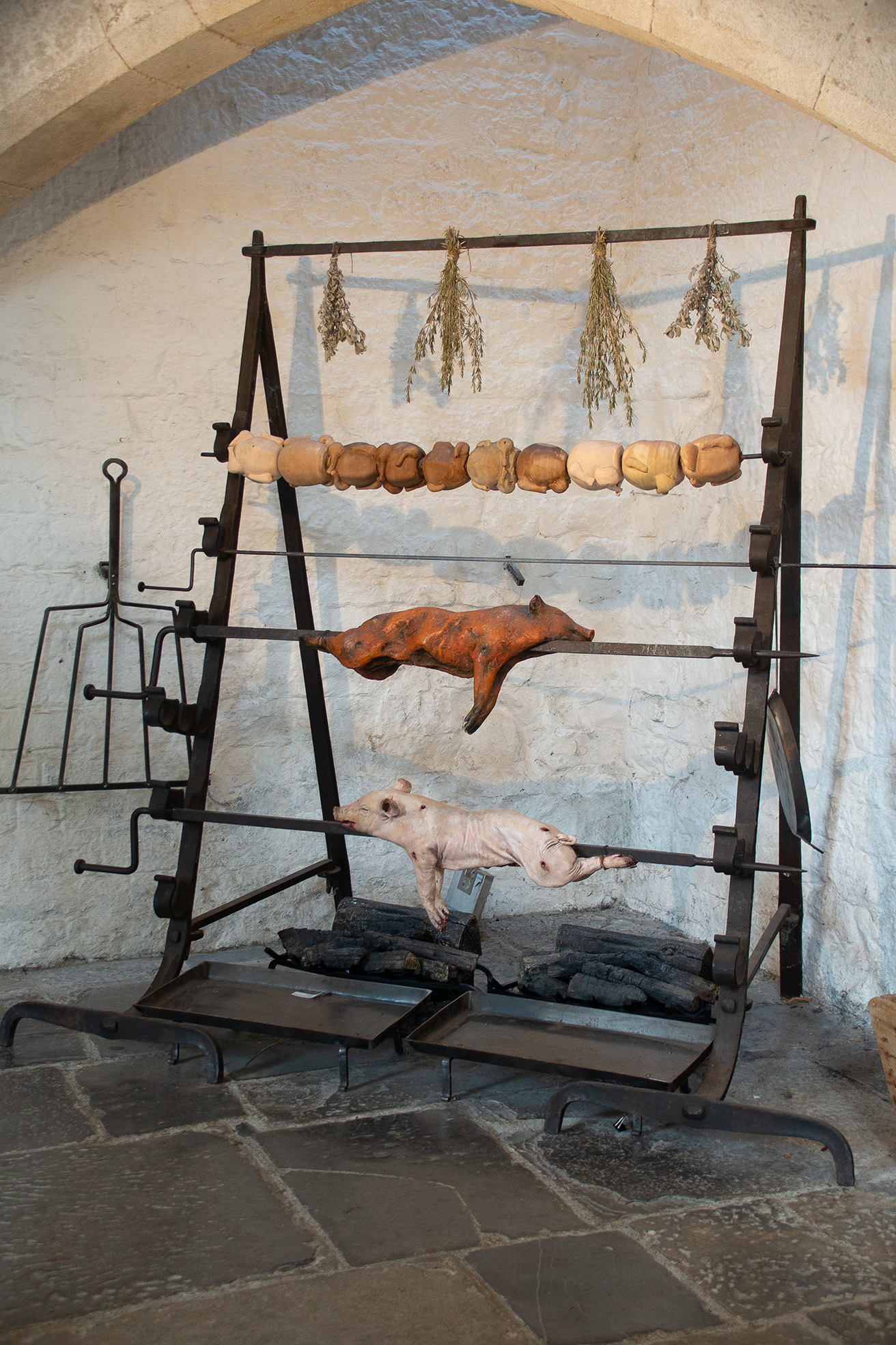 The image depicts a traditional roasting setup with a pig and a boar being roasted on a spit over an open fire. Above them, there are various vegetables and herbs hanging, likely for drying or as part of the roasting process. The setup is situated in a rustic, stone-walled room, suggesting a historical or medieval cooking method.