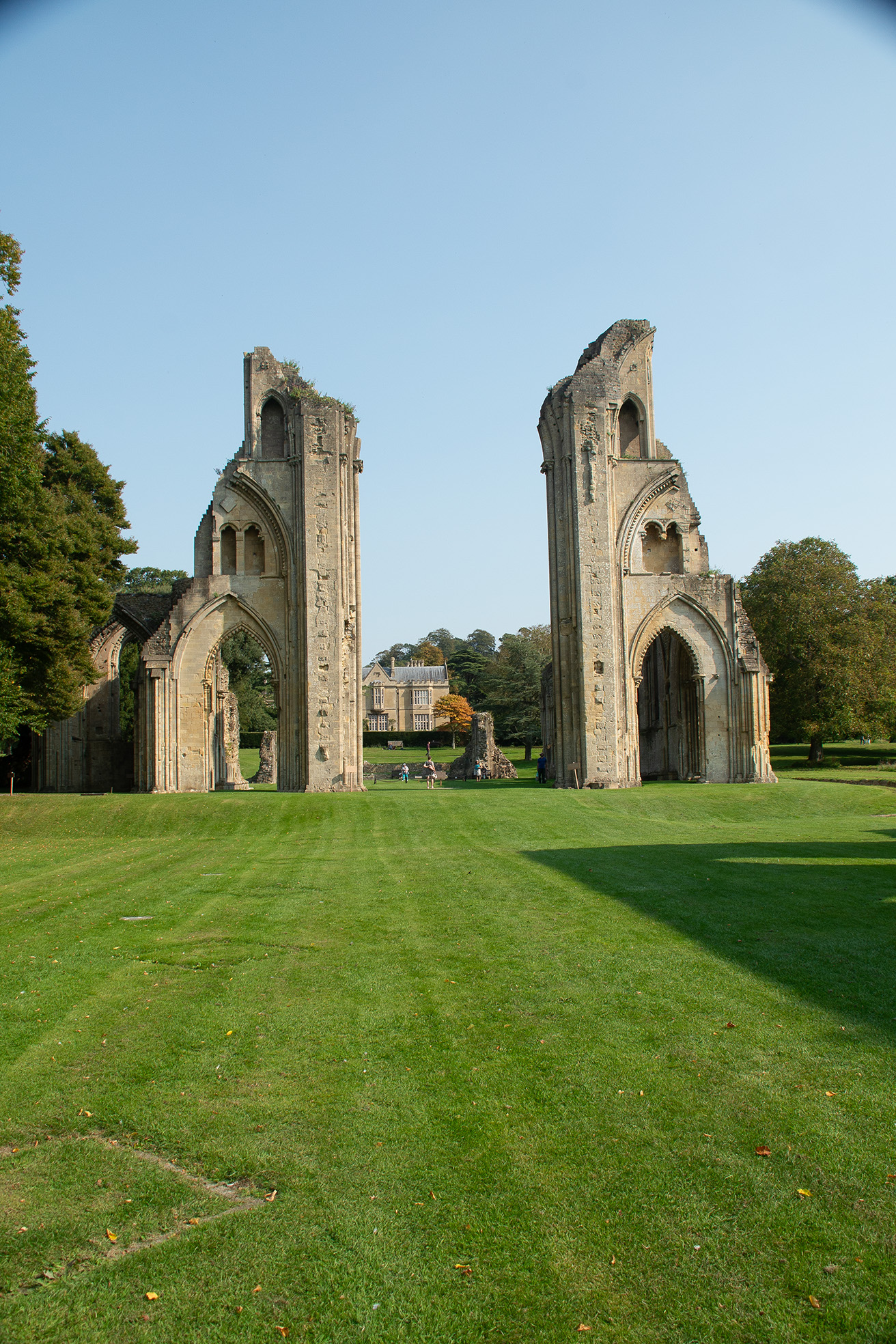 The image depicts the ruins of a historic abbey or cathedral, characterized by its tall, weathered stone arches and remnants of walls. The scene is set in a well-maintained grassy area with a clear blue sky overhead. In the background, a large, stately house is visible, surrounded by trees. The overall atmosphere is serene and picturesque, suggesting a Brassknocker of historical significance and beauty.