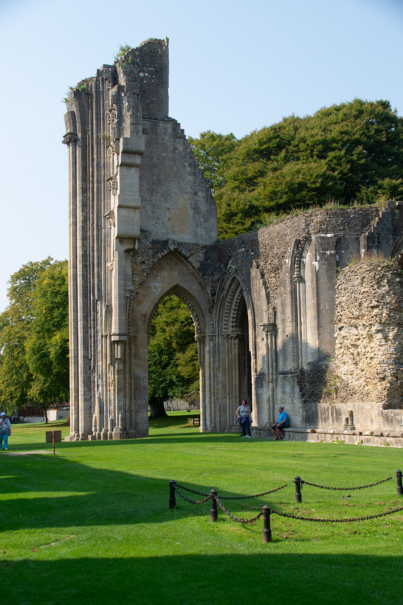 The image depicts the ruins of an old stone building, likely a historical or medieval structure, with tall, arched windows and intricate stonework. The ruins are set in a grassy area with a few people nearby, suggesting it is a tourist site or place of interest. The surrounding area is lush with greenery, and the sky is clear, indicating a sunny day. The overall scene conveys a sense of history and tranquility.