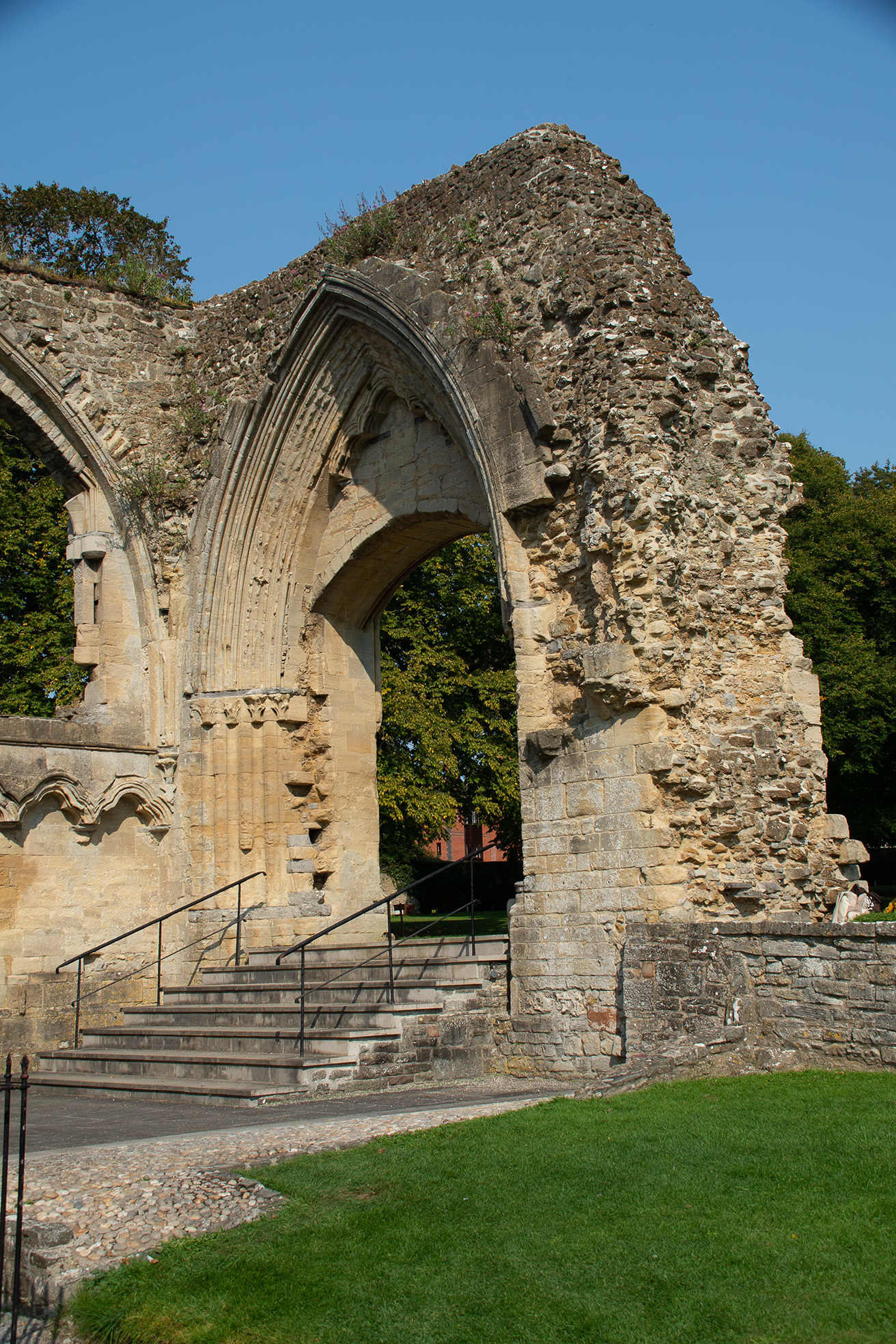 The image depicts the ruins of an old stone building, likely a historical or medieval structure. The remnants include a large, intricately designed archway with detailed stone carvings, supported by partially standing walls. There are steps leading up to the archway, and the area around the ruins is grassy with a clear, sunny sky in the background.