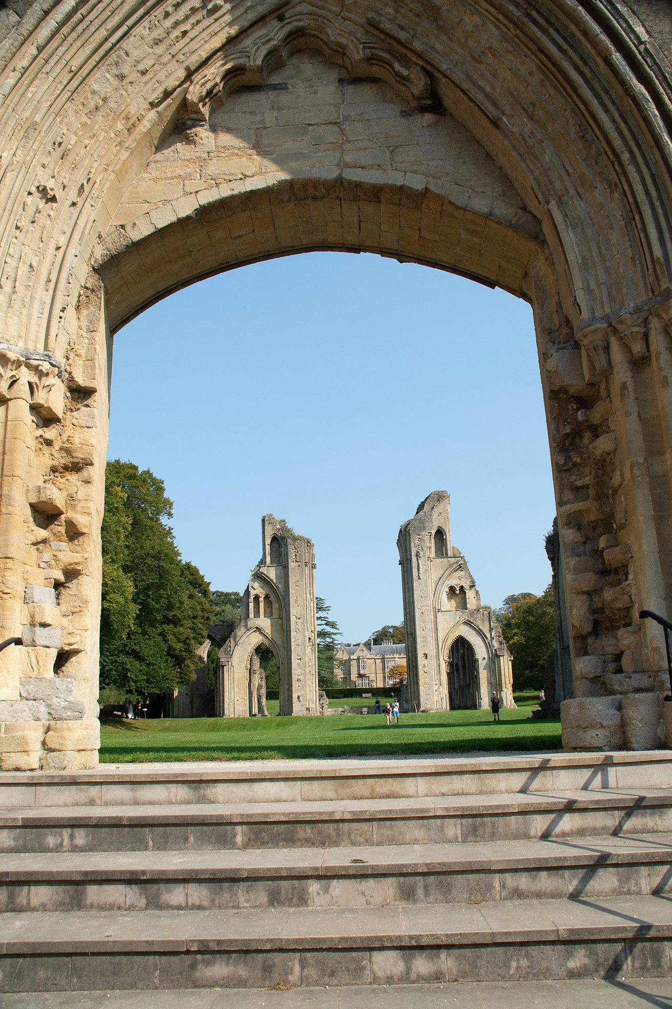 The image depicts the ruins of an old stone structure, possibly a historical or medieval building, viewed through a large archway. The ruins consist of tall, partially collapsed walls and arches, set within a well-maintained grassy area. The sky is clear and blue, and there are a few people walking and standing around the ruins, suggesting it is a tourist site or a place of historical interest.