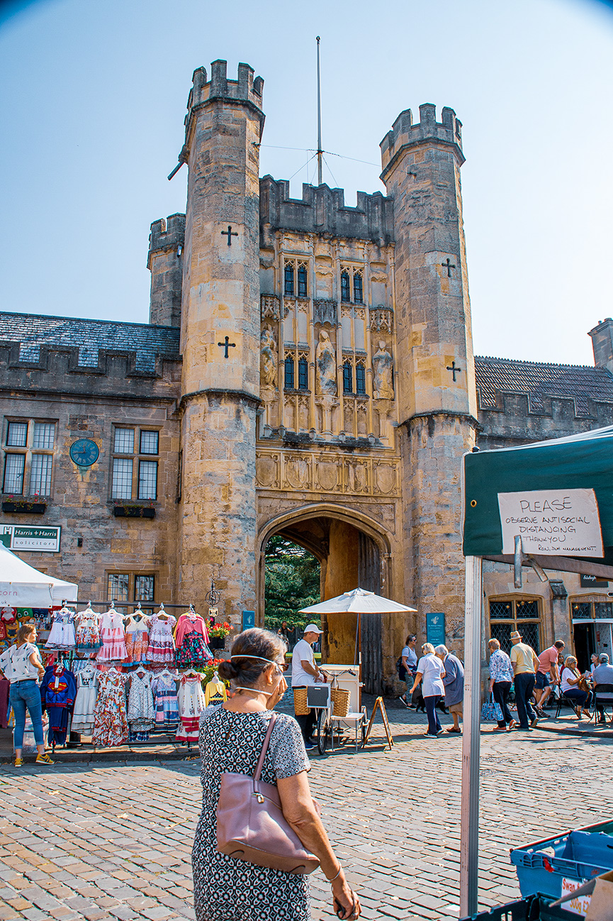 The image depicts a bustling scene in front of a historic castle gatehouse. The gatehouse features two large towers with crosses on them and intricate stone carvings above the archway. Various people are seen walking around, shopping at stalls selling colorful clothing, and sitting at outdoor tables. A sign asks visitors to observe social distancing. The overall atmosphere is lively and vibrant, with a mix of tourists and locals enjoying the area.