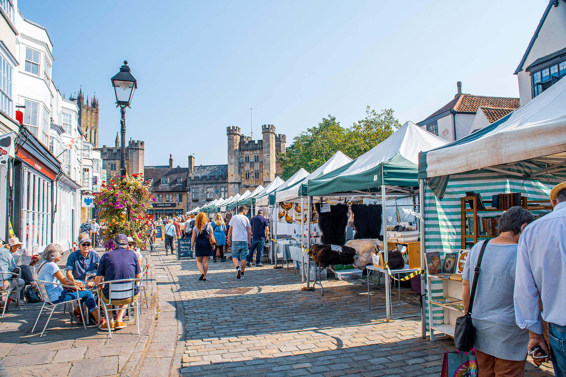 The image depicts a bustling outdoor market set in a charming, historic town. The market features numerous stalls with green and white canopies displaying various goods, including artwork and textiles. People are seen walking, browsing, and sitting at outdoor cafes along the cobblestone street. The architecture includes a mix of modern and historic buildings, with a prominent castle-like structure in the background. The scene is lively and vibrant, with clear skies and a sunny atmosphere.