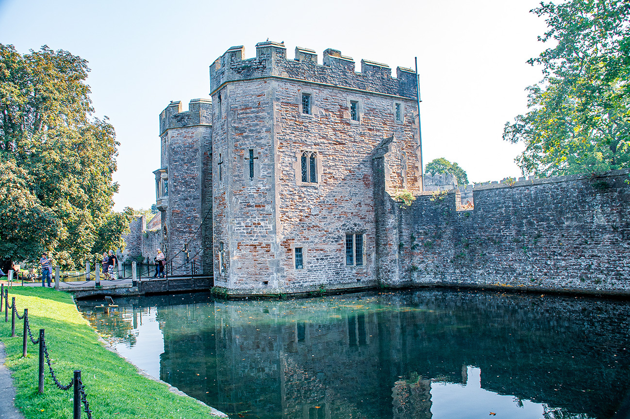 The image depicts a historic castle with stone walls and a moat, surrounded by greenery and trees. The castle features multiple towers with battlements and small windows. People are seen walking along a pathway near the castle, and the scene is reflected in the calm water of the moat.