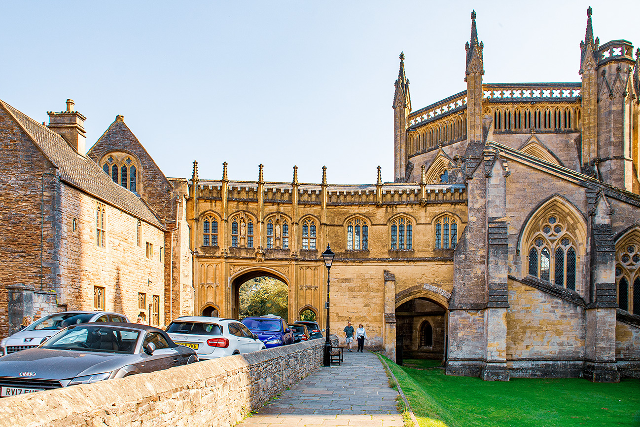 The image depicts an architectural scene featuring a historic building with Gothic elements, such as pointed arches, intricate stonework, and tall spires. The building appears to be part of a larger complex, possibly a cathedral or university. Cars are parked along the side of a narrow road leading to an arched gateway. The setting is well-maintained, with a stone wall and a small patch of green grass. The overall atmosphere is serene and picturesque, suggesting a location of historical and cultural significance.