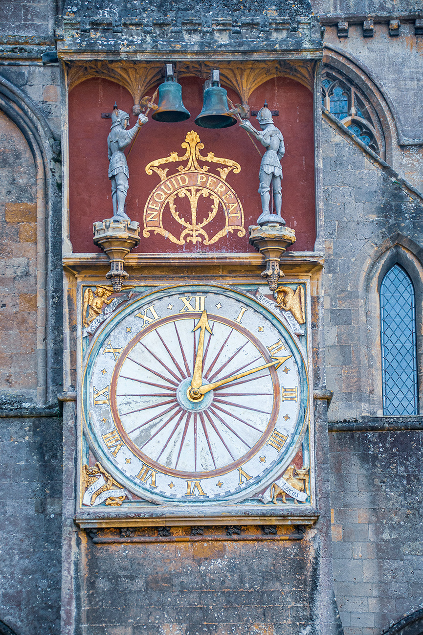 The image depicts an ornate astronomical clock with Roman numerals and intricate details, including two statues ringing bells and a Latin inscription. The clock is part of a historic building with Gothic architectural elements.