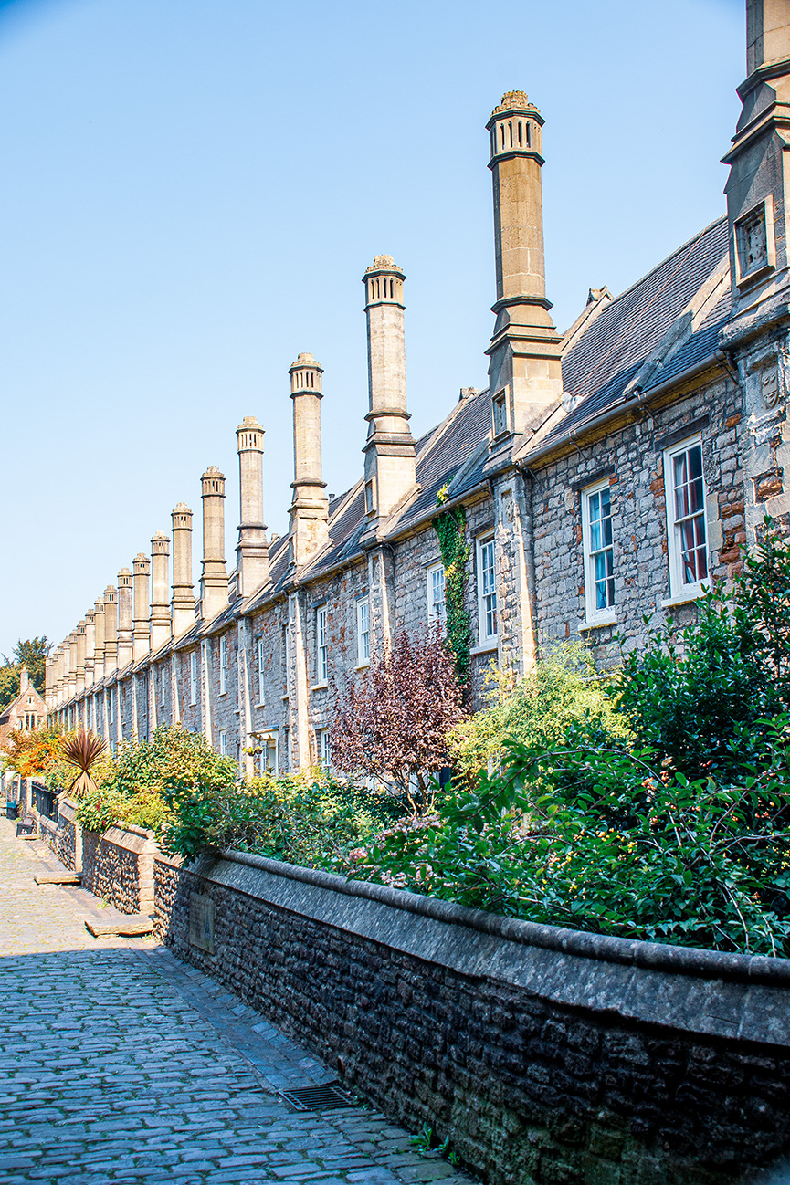 The image depicts a row of historic stone buildings with tall, ornate chimneys, likely part of a university or college. The buildings have a traditional architectural style with slate roofs and stone walls. The scene is set on a cobblestone path bordered by a stone wall and lush greenery, suggesting a serene and picturesque environment.