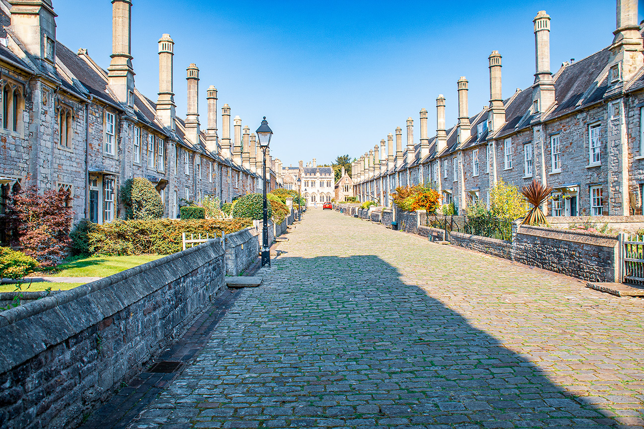 The image depicts a picturesque street in what appears to be a historic or traditional European town. The street is lined with uniform, stone-built buildings featuring numerous chimneys and small windows. The cobblestone path in the center is flanked by well-maintained gardens with various plants and shrubs. The scene is bathed in bright sunlight, creating a serene and inviting atmosphere.