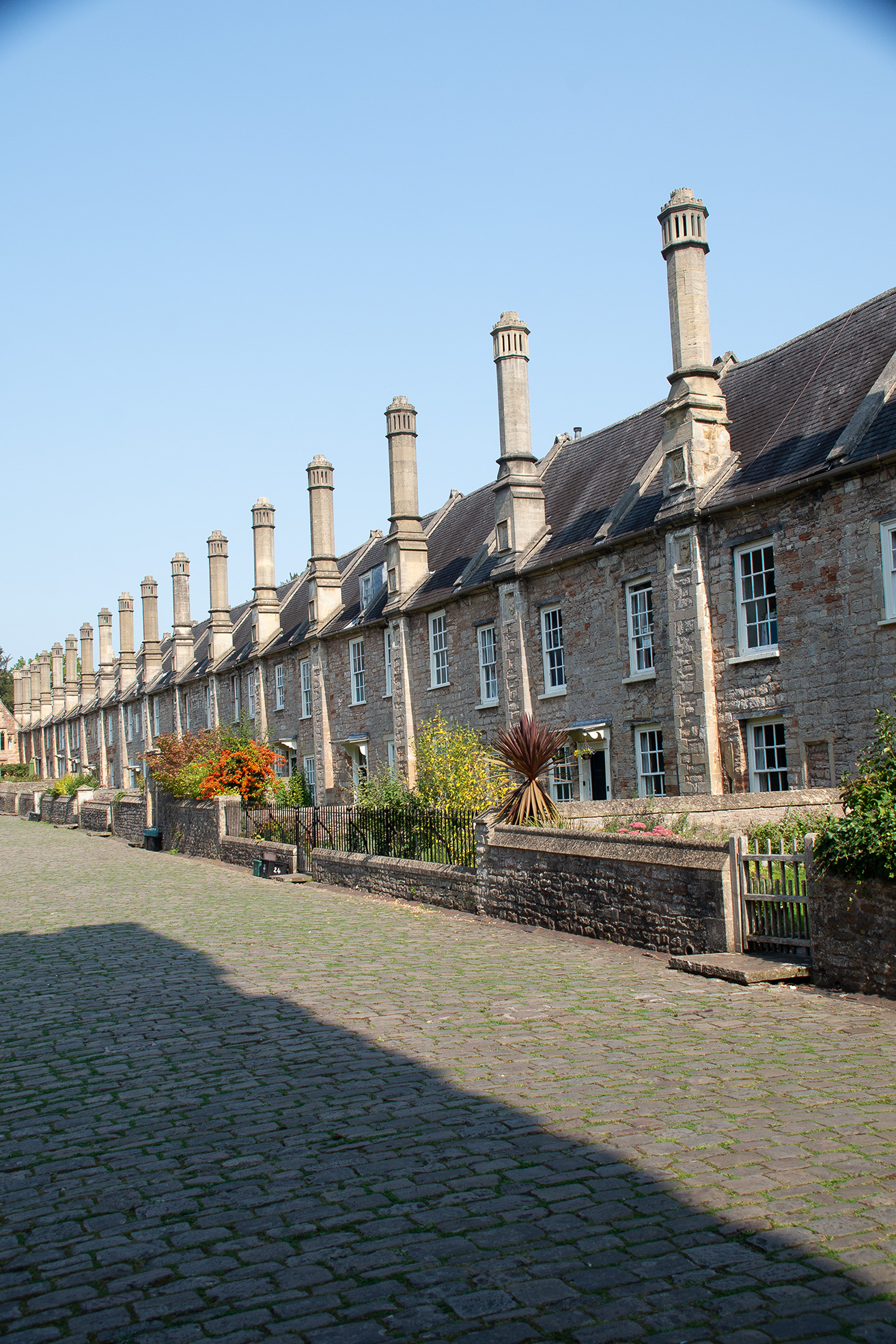 The image depicts a row of historic, stone-built houses with distinctive chimneys, likely from the 17th or 18th century. The houses have a uniform architectural style, featuring large windows and steeply pitched roofs. The street in front is paved with cobblestones, and there are small, well-maintained gardens with plants and flowers in front of each house. The overall scene suggests a quaint, picturesque setting, possibly in a rural or small-town environment.
