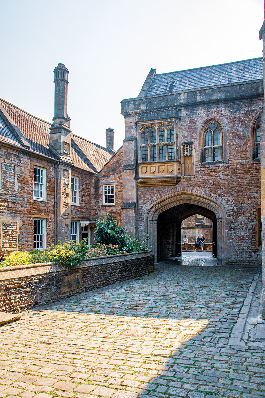 The image depicts a historic building with a cobblestone courtyard. The architecture features a mix of brick and stone, with large windows and a prominent archway. There are two people visible through the archway, and the building has several chimneys and a tiled roof. The courtyard is lined with plants and flowers, adding a touch of greenery to the scene.