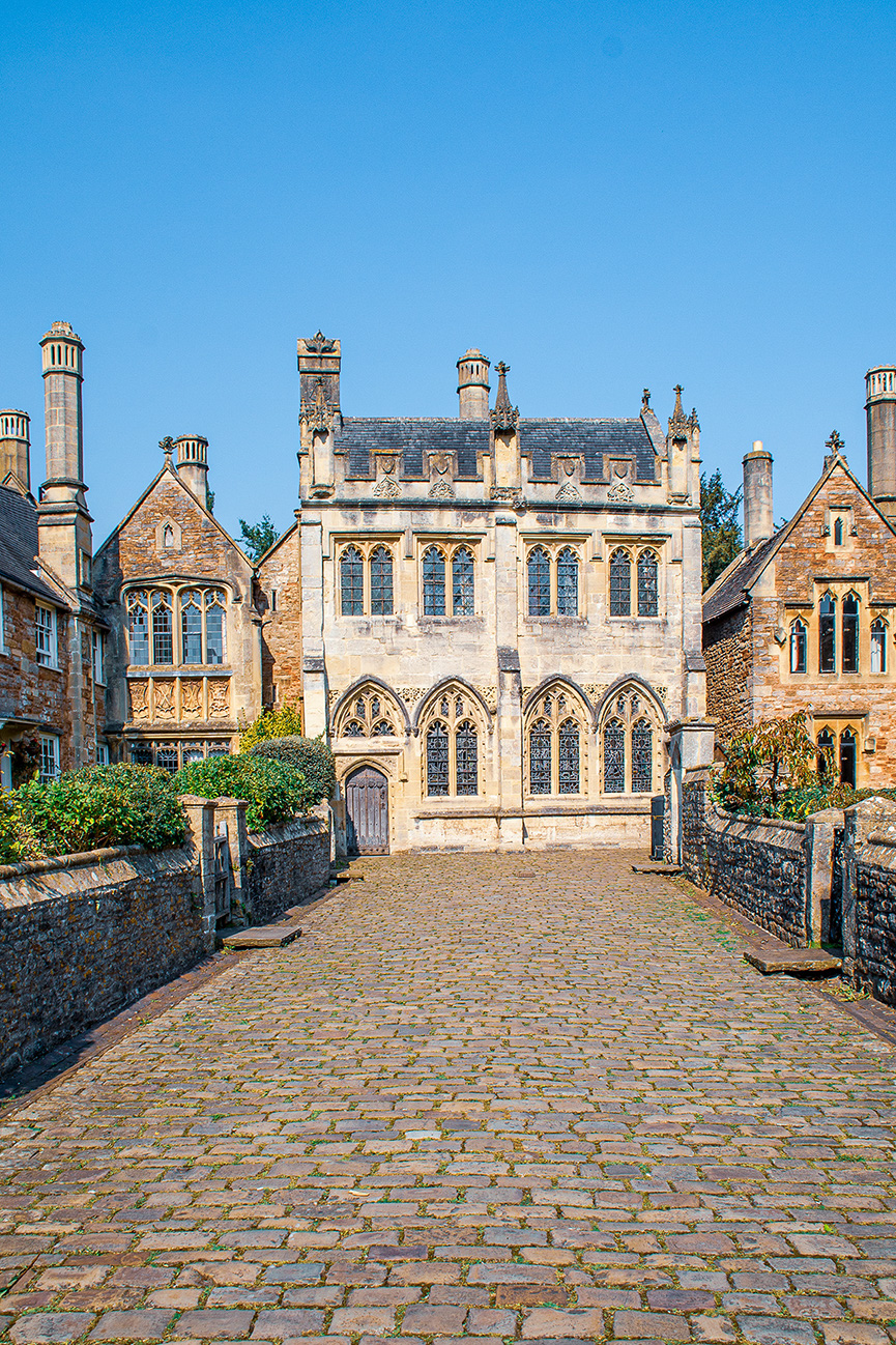 The image depicts a historic building with Gothic architectural elements, such as pointed arches and intricate stonework. The building is surrounded by a cobblestone courtyard with low stone walls and some greenery. The sky is clear and blue, suggesting a sunny day.